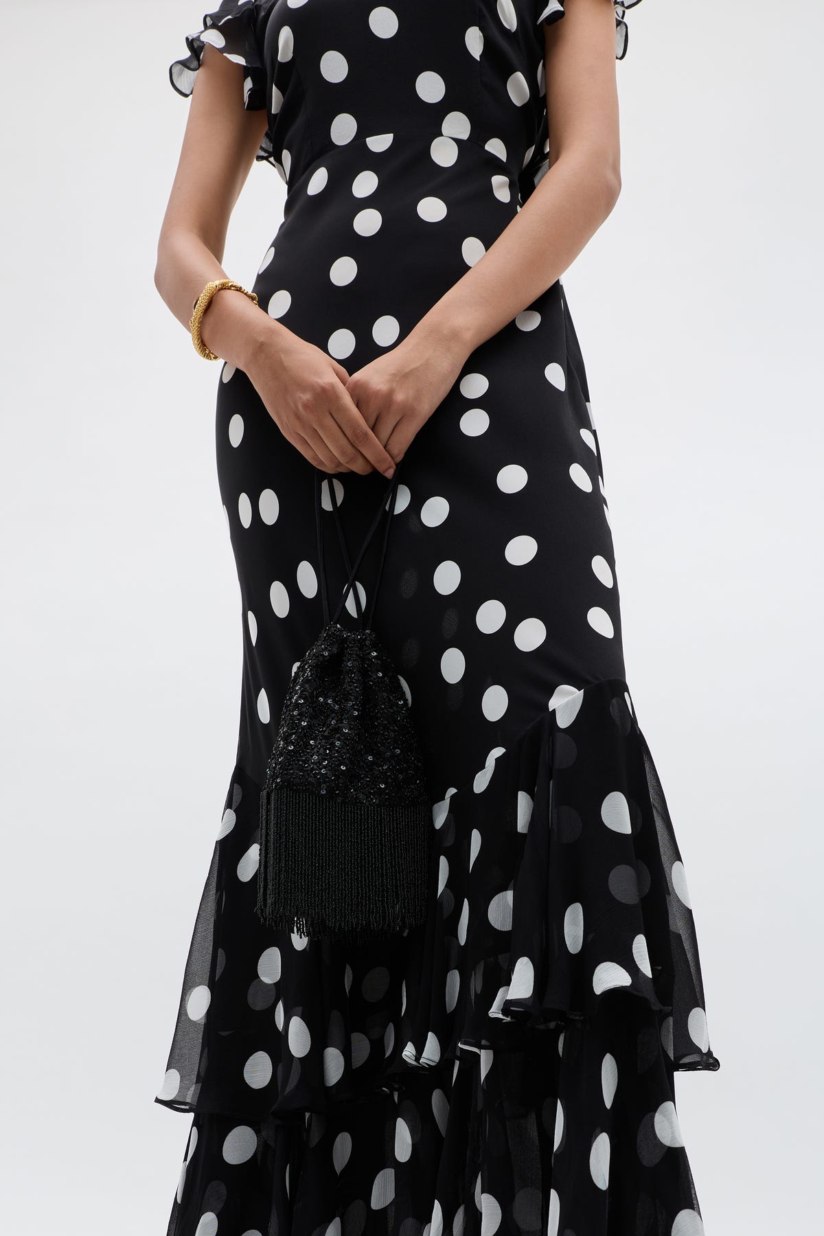 A person wearing the Ordinia - Mono Polka Dot Black dress, featuring vintage-inspired white polka dots and a ruffled hem, holds a black beaded fringe purse. Only their torso and arms show against a plain light background.