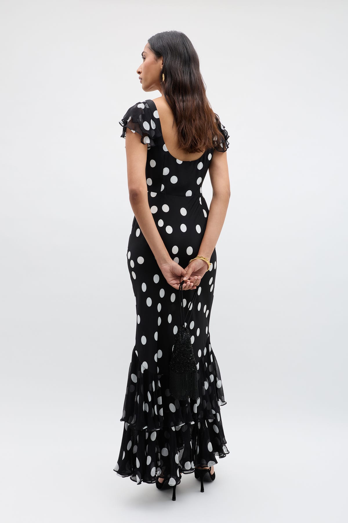 A woman stands facing away in the Ordinia Mono Polka Dot Black gown, featuring vintage-inspired white dots and a ruffled hem. She holds a black clutch behind her back, with wavy hair down and black heels against a plain background.