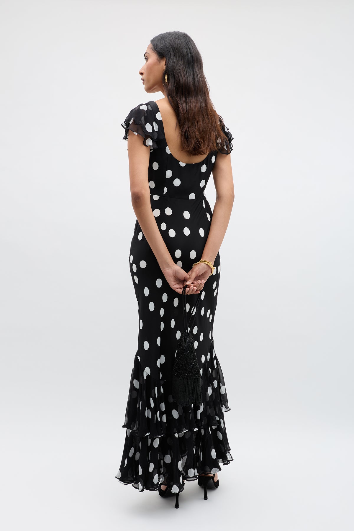 A woman stands facing away in the Ordinia Mono Polka Dot Black gown, featuring vintage-inspired white dots and a ruffled hem. She holds a black clutch behind her back, with wavy hair down and black heels against a plain background.