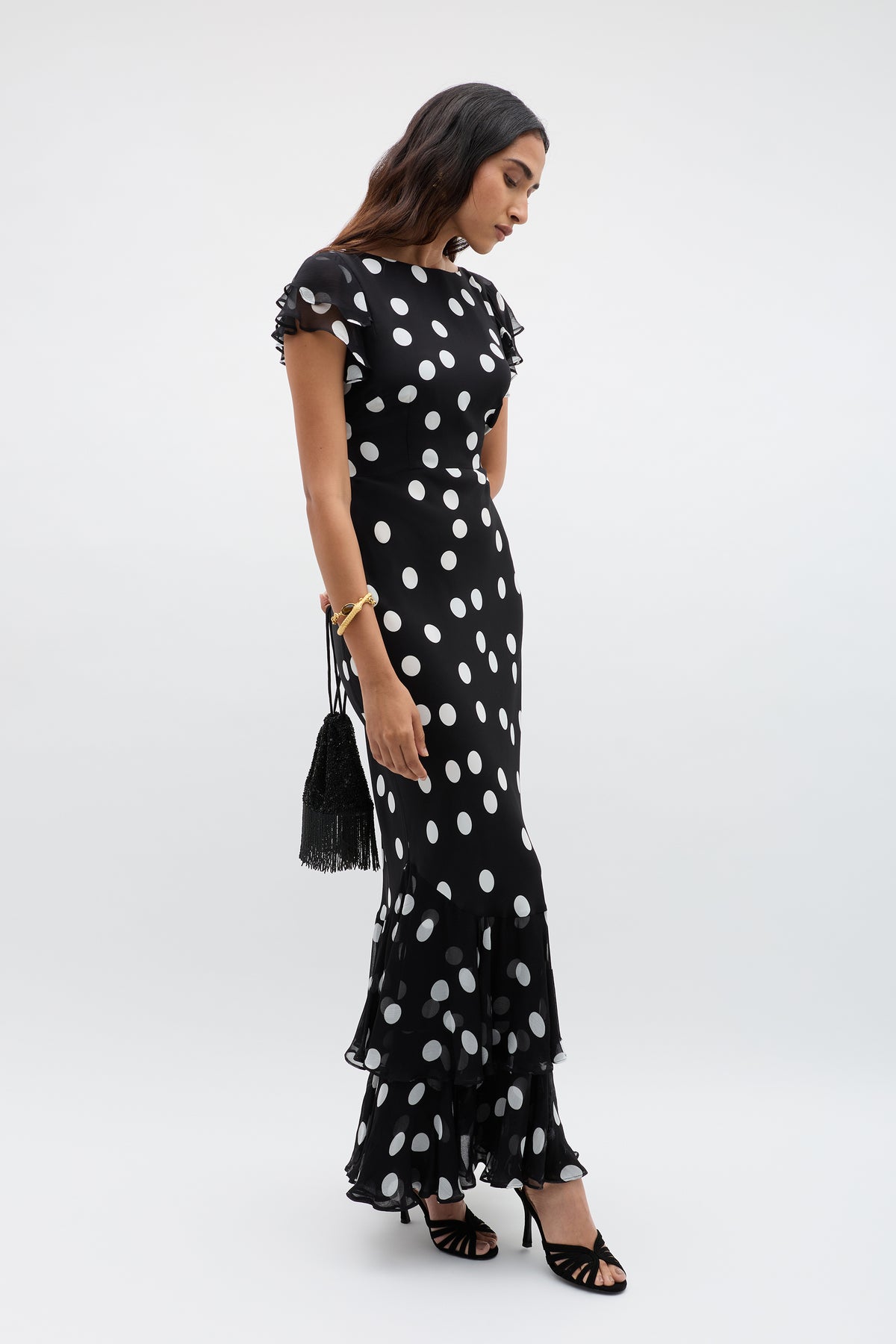 Wearing the Ordinia - Mono Polka Dot Black dress, a woman with a ruffled hem and short sleeves looks down. She accessorizes with a black tassel handbag, black heels, and a gold bracelet.