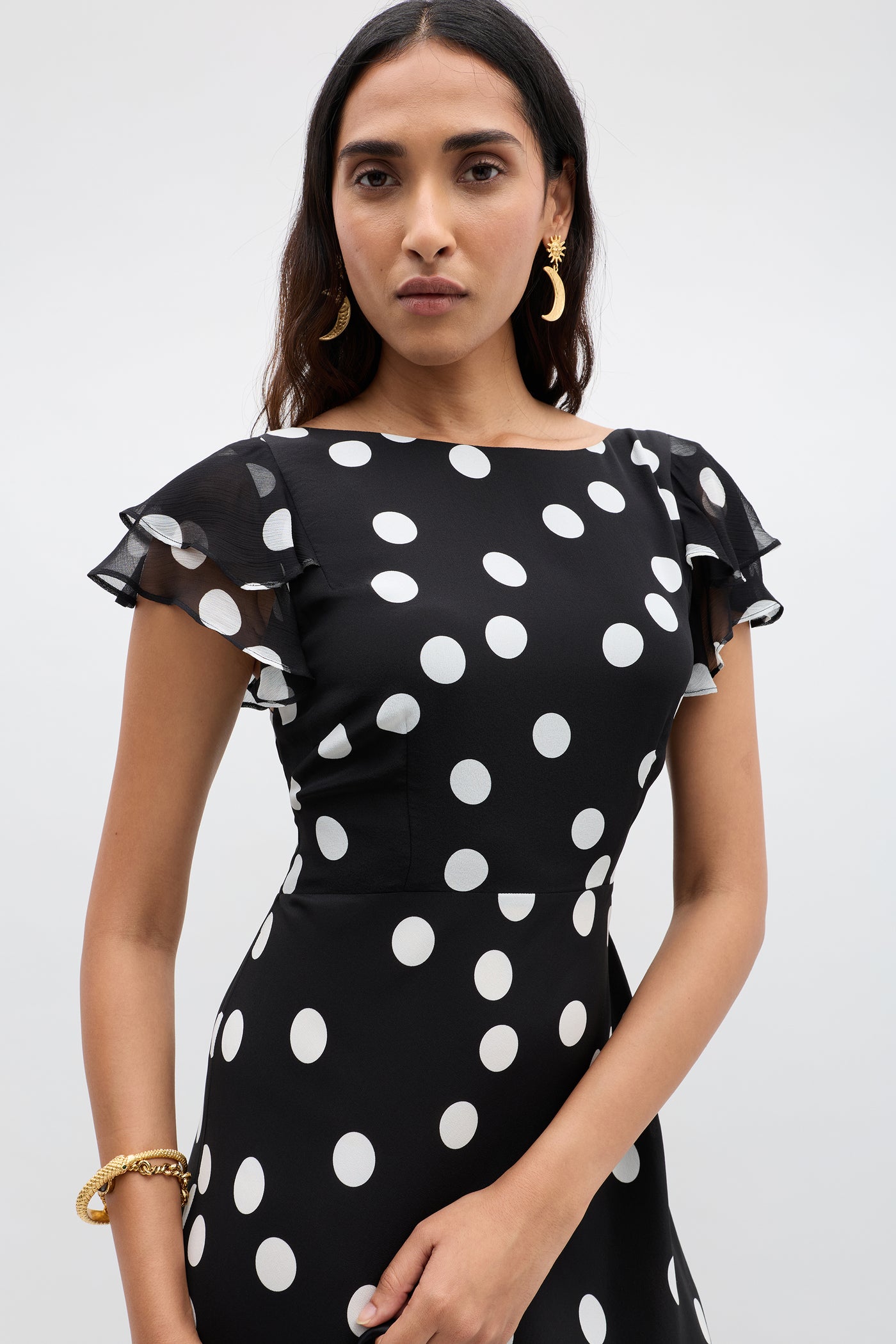 A woman in the Ordinia Mono Polka Dot Black dress, featuring white dots and ruffled short sleeves, poses against a plain light background. She wears gold earrings and a bracelet, with long dark hair and a neutral expression.