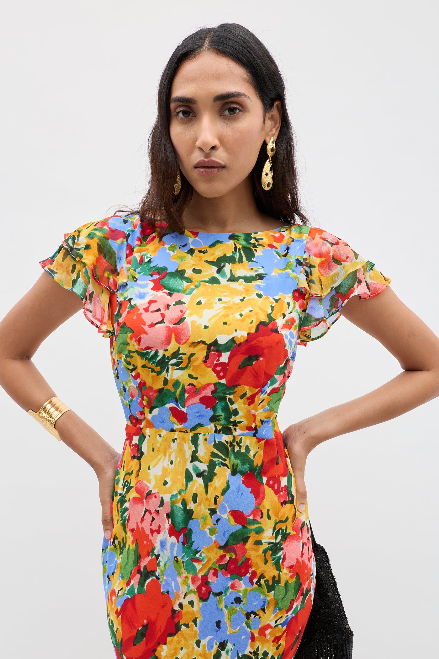 A woman with long dark hair wears the Ordinia - Wedding Floral Multi dress with ruffled short sleeves, gold drop earrings, and a bracelet, standing against a plain background—ideal for formal occasions.