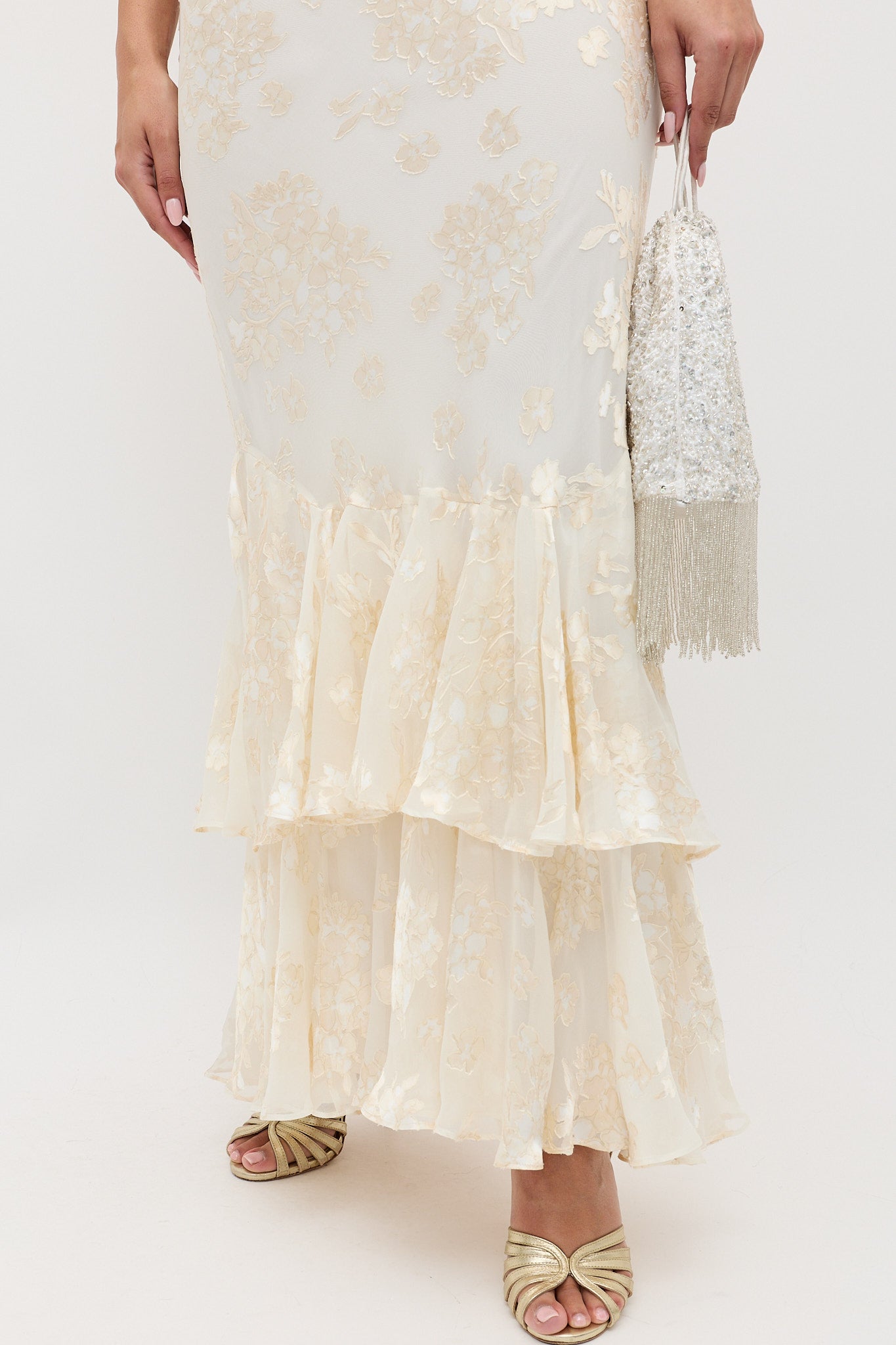 A woman wears the Ordinia - Bouquet Devore Cream floral ruffled skirt, holding a silver beaded fringe bag and wearing gold open-toe heels against a plain white background.