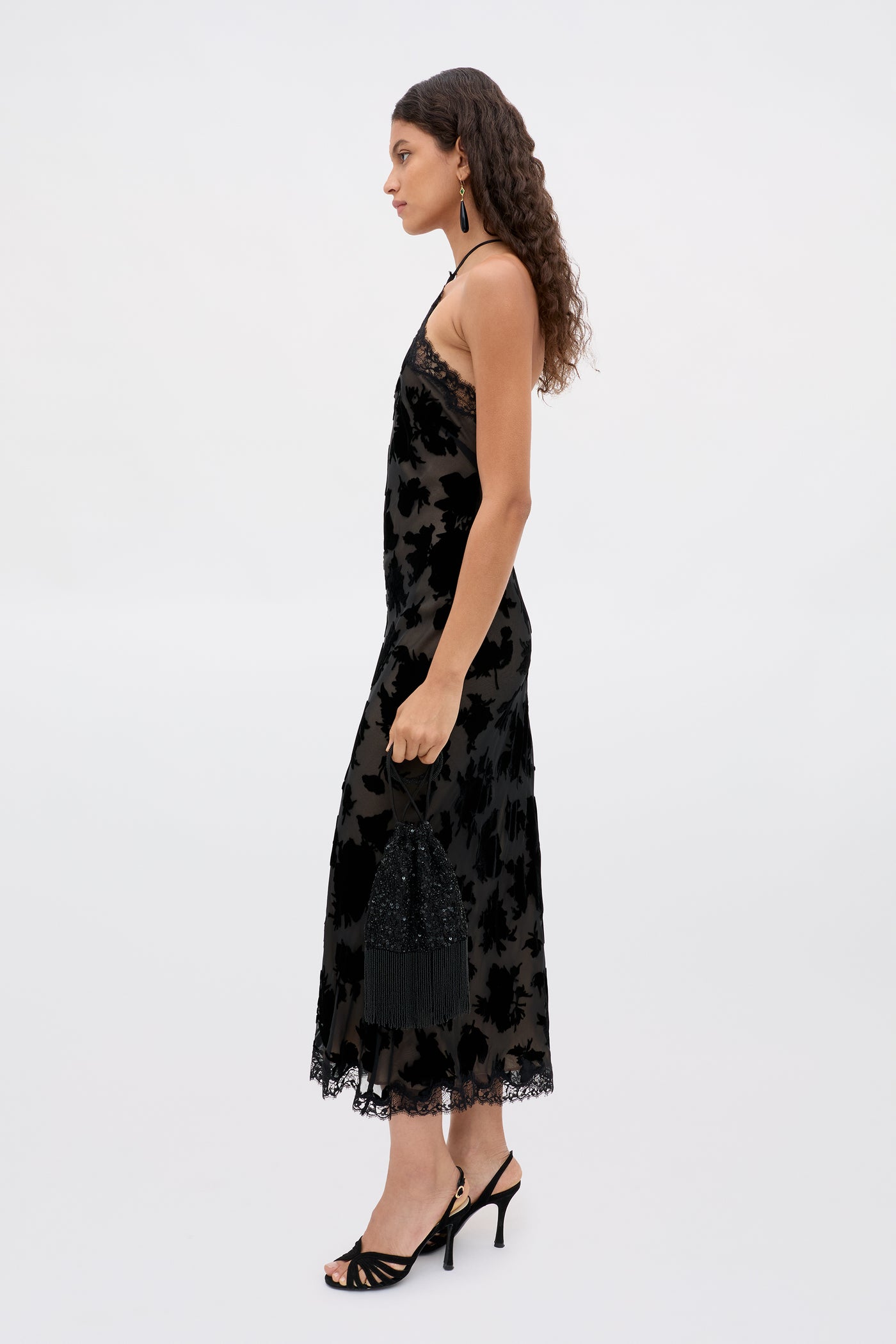 A woman with long curly hair, in a sleeveless black lace Harlan - Countryside Devore Mono dress and black heels, stands in profile against a plain light background, holding a small black fringed handbag.