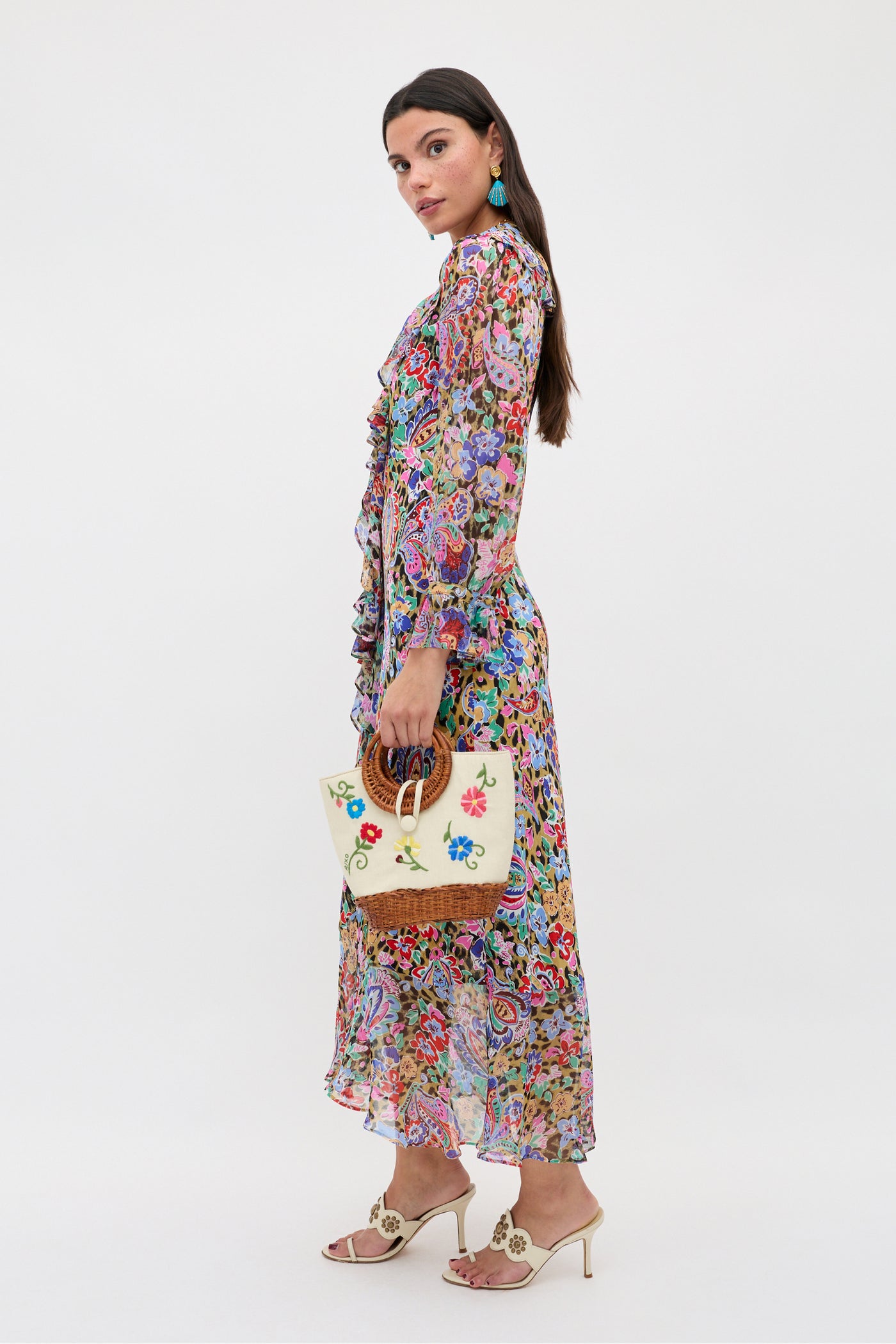A woman in the Gracye - Cara Floral Leopard Brown long, sheer dress and cream heeled sandals stands sideways, holding a decorative handbag. She has long dark hair and turquoise earrings against a plain white background.