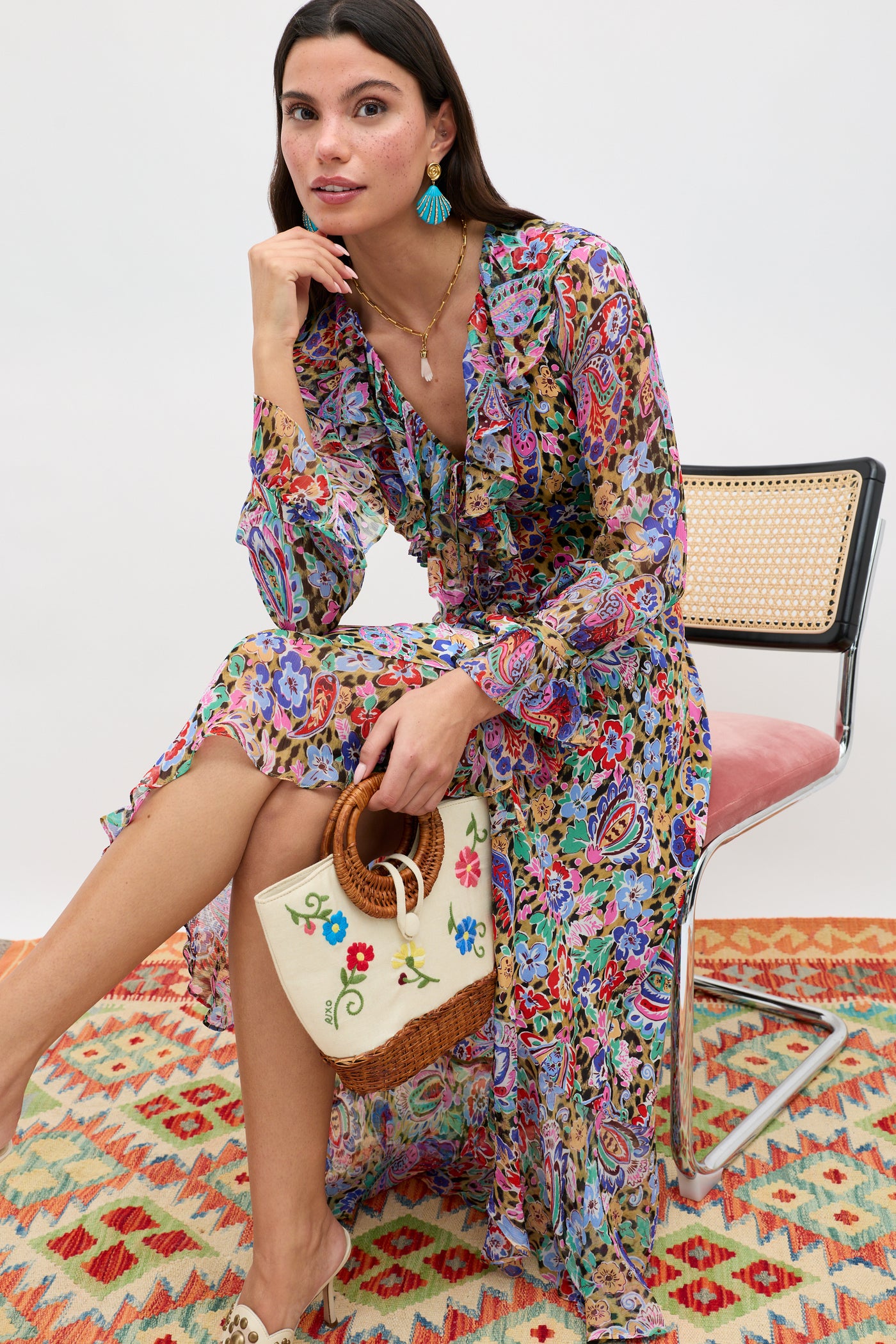A woman in a long, colorful floral dress sits on a woven chair, holding the Zelene - Ombre Floral Embroidery Cream bag with a wicker handle. She wears turquoise earrings, a necklace, open-toe shoes, and sits atop a patterned rug.