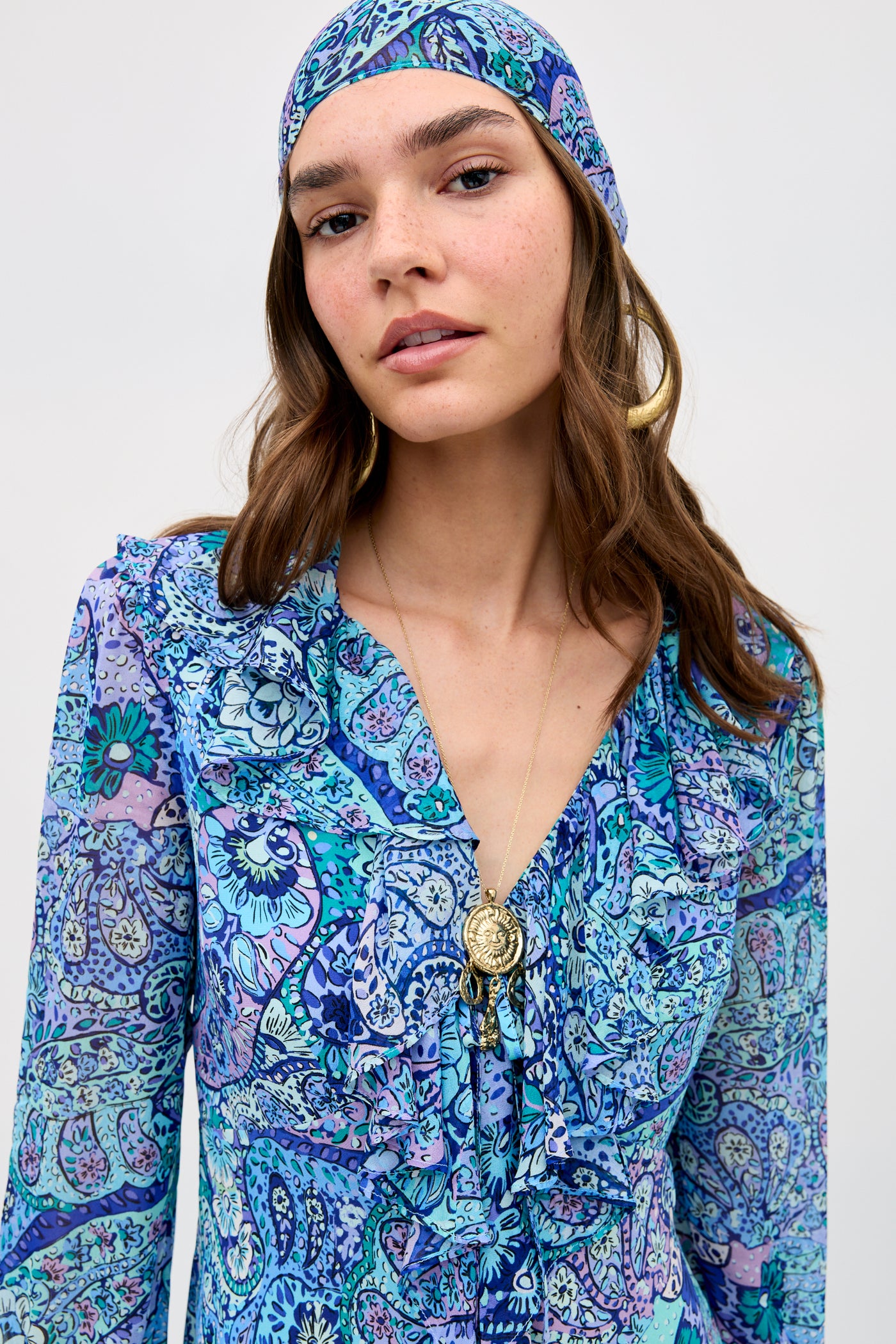 A woman wearing the Julien - Paisley Floral Blue dress with a matching headscarf, bold earrings, and a long pendant necklace stands confidently against a neutral background.
