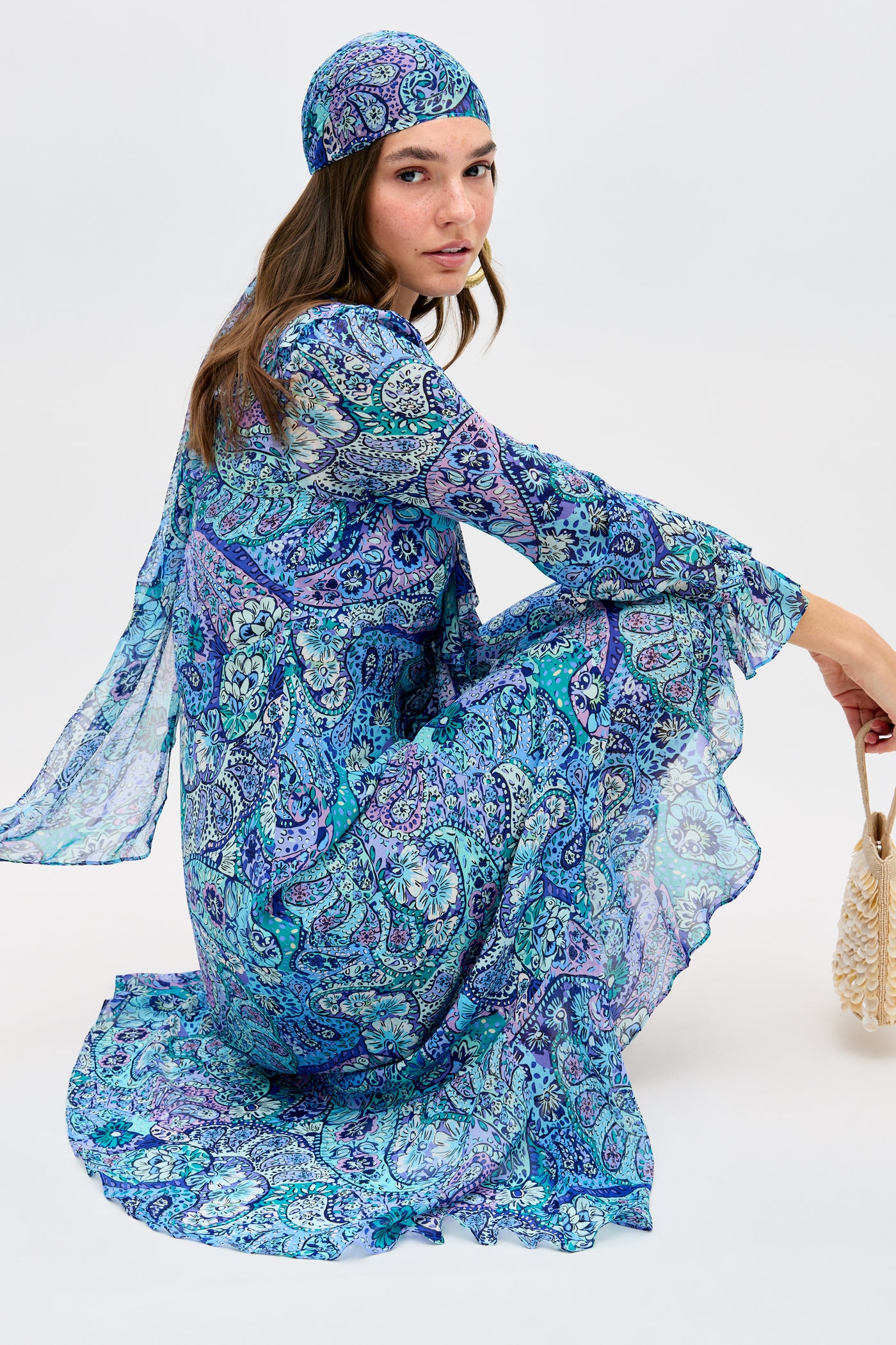 A woman sits sideways on the ground, wearing the Julien - Paisley Floral Blue long-sleeved dress with a matching headscarf. She holds a small woven bag and looks over her shoulder.