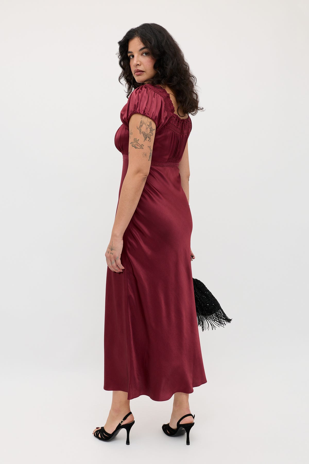 A woman with curly dark hair wears the Maeva - Maroon dress and black heels, holding a black fringed bag. Tattooed on her arm, she stands against a plain white background, glancing over her shoulder.