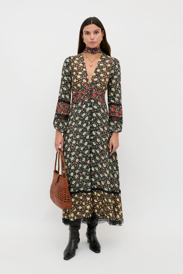A woman stands against a white background wearing the RIXO Raven - Eva Floral Mix Black dress with long sleeves, a deep V-neck, and red floral accents. She pairs it with a brown woven bag, black boots, loose hair, and minimal accessories.