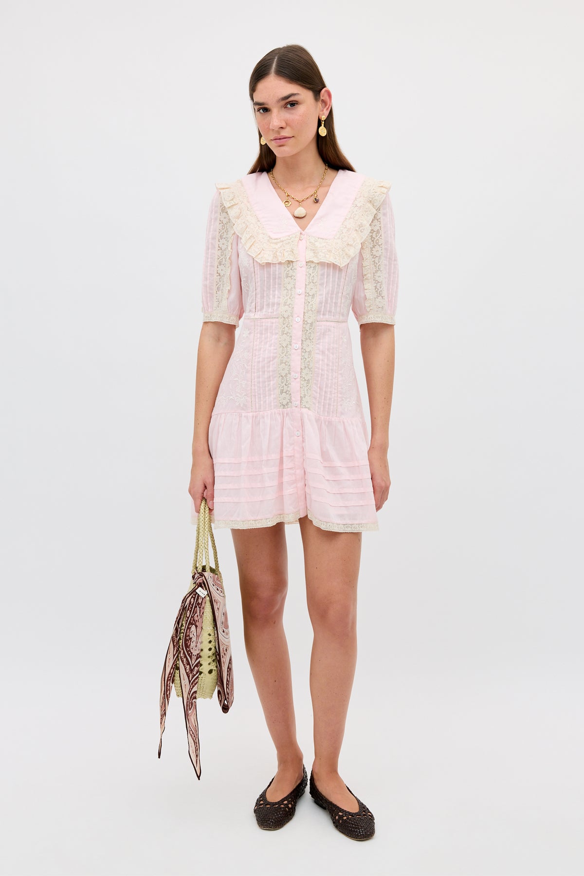 A woman poses against a plain background in the Dove - Pansy Embroidery Vintage Pink dress, which features lace embroidery and a tiered skirt. She accessorizes with a patterned bag, dark ballet flats, and gold earrings for an elegant look.