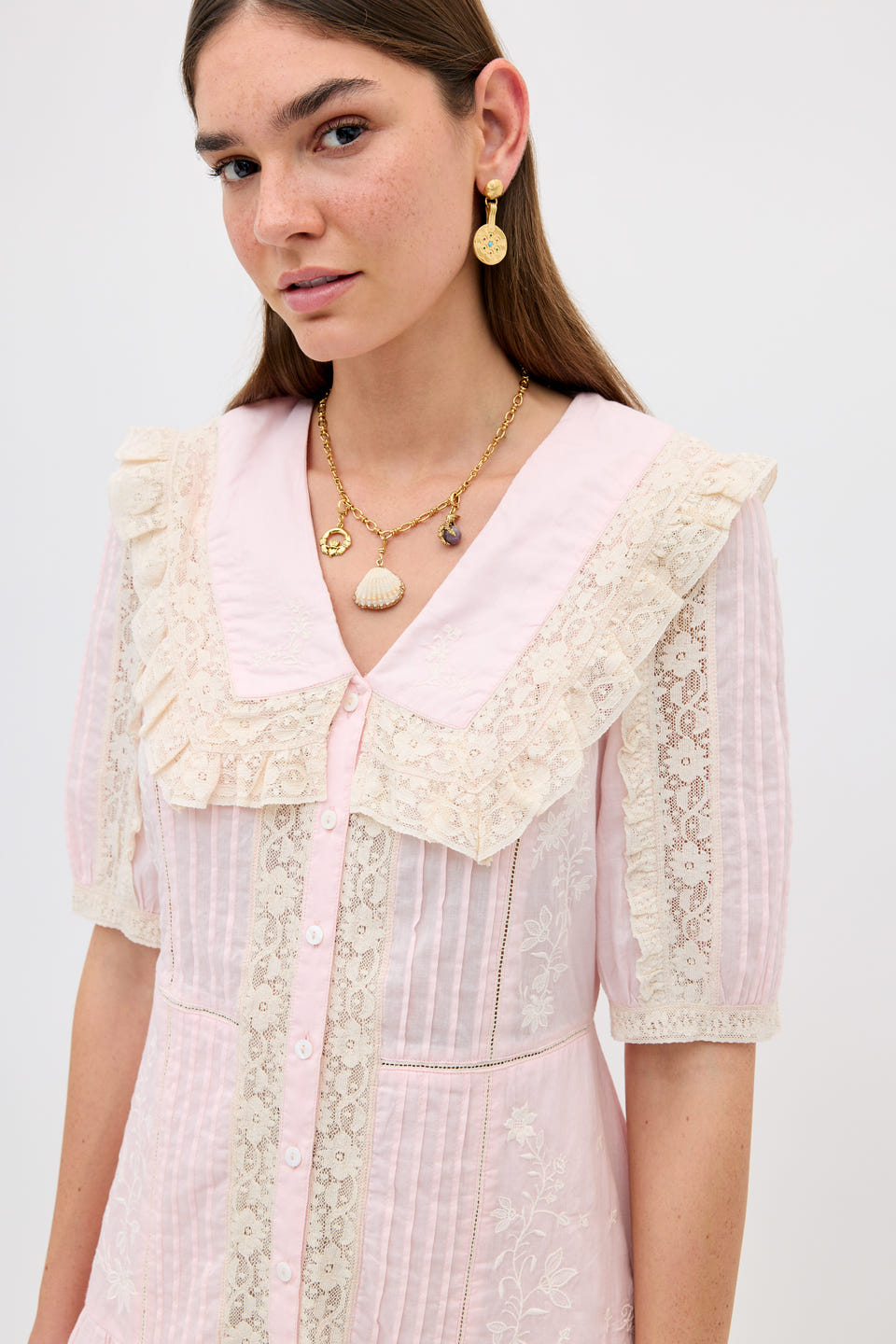 A woman in a light pink dress with lace and ruffles wears gold earrings, layered necklaces, and the Orsina - Gold pendant inspired by traditional Irish jewelry, standing against a plain light background.
