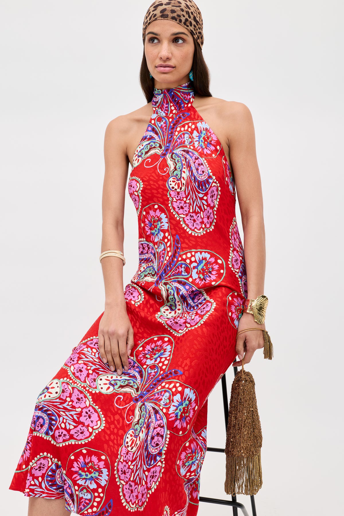 A woman in the Maura - Butterfly Bloom Jacquard Red dress sits on a stool, accessorized with a leopard print headscarf, gold bracelets, and a brown fringe handbag against a plain background.