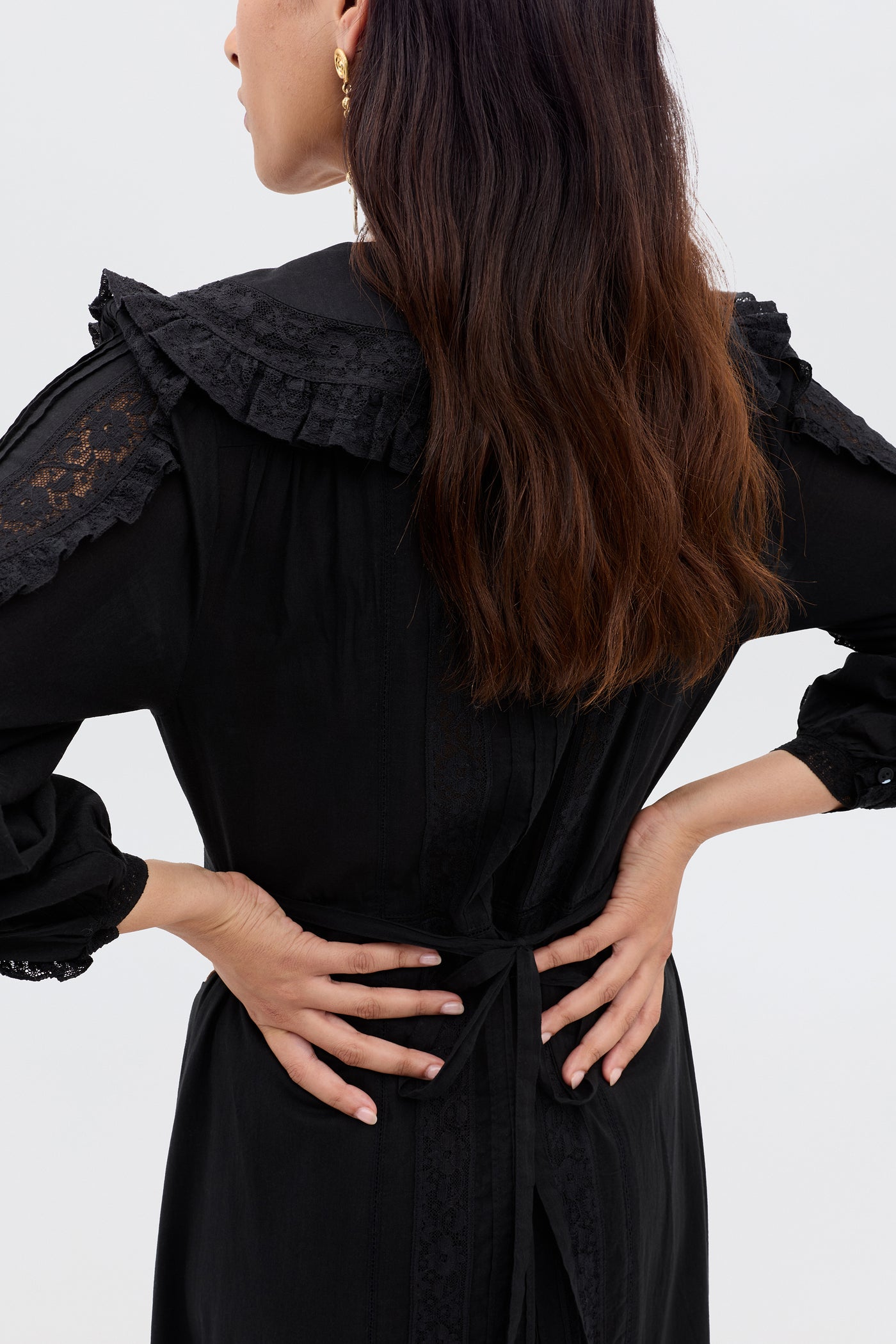 A woman with wavy brown hair stands with her back to the camera, wearing the Hilary - Black dress with lace details and a ruffled collar. Her hands rest on her hips against a plain light background.