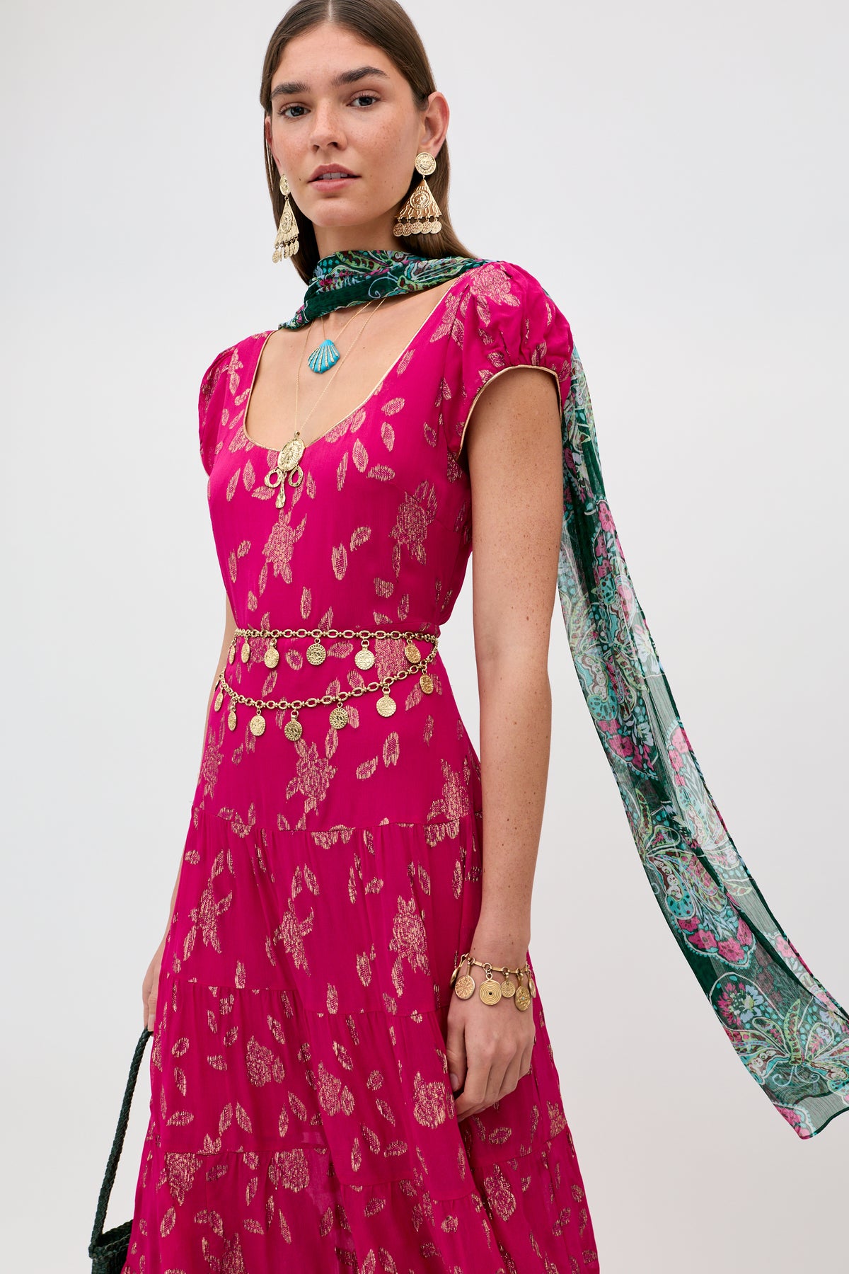A woman wears the Lita - Gold dress, featuring gold patterns and layered gold jewelry, accessorized with a green patterned scarf, standing against a plain light background.