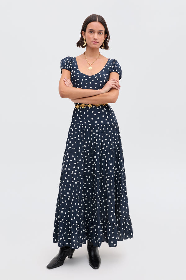 A woman stands with arms crossed, wearing the Remi - Bohemia Spot Deep Navy dress with a black belt and boots, hoop earrings, and a gold necklace against a plain background.