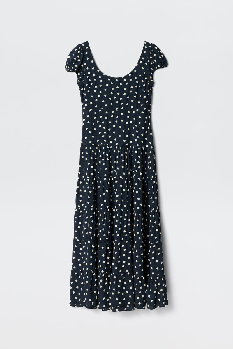 The Remi - Bohemia Spot Deep Navy dress features a scoop neck, white polka dots, short sleeves, and an ankle-length tiered skirt—ideal for summer and shown on a plain white background.