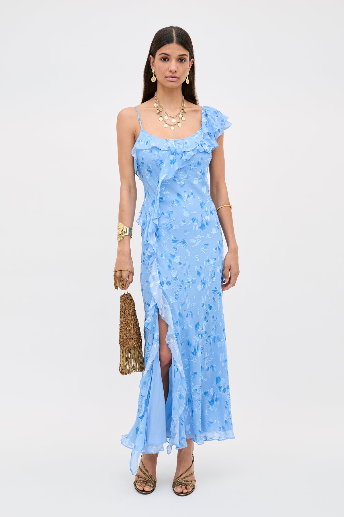 A woman stands against a plain light background wearing the Ester - Ava Devore Blue floral, ruffle-trimmed one-shoulder dress with a thigh-high slit, gold jewelry, brown heeled sandals, and a gold tasseled handbag.