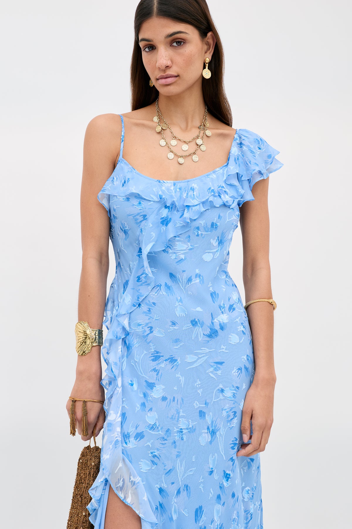 Wearing the Ester - Ava Devore Blue dress, a woman stands against a plain background. She accessorizes with layered gold jewelry, a gold bracelet, and carries a brown textured handbag.