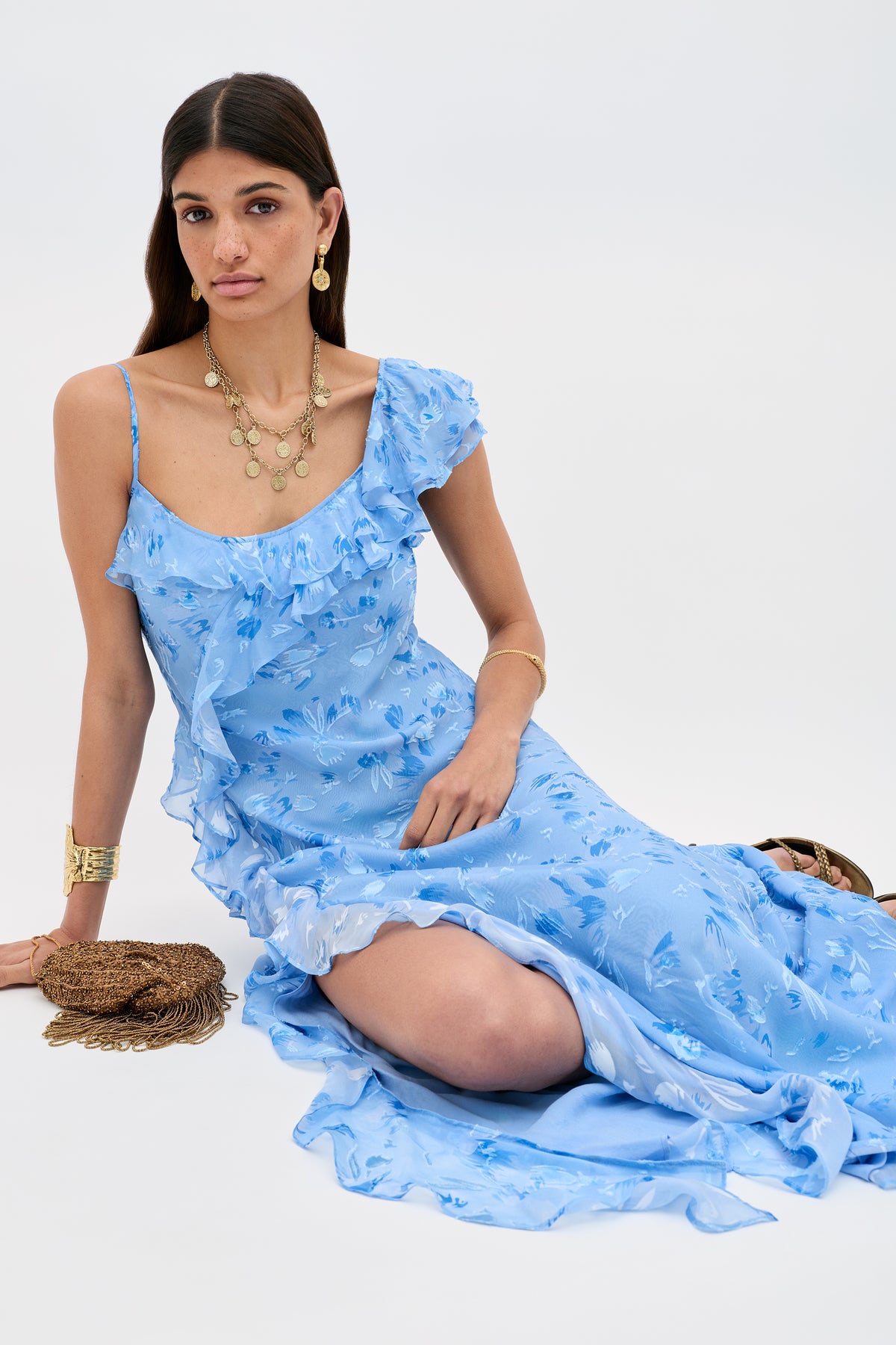 A woman with long brown hair sits on the floor wearing the Ester - Ava Devore Blue dress, paired with gold earrings, layered necklaces, a bracelet, and a brown woven handbag. The background is plain and light colored.