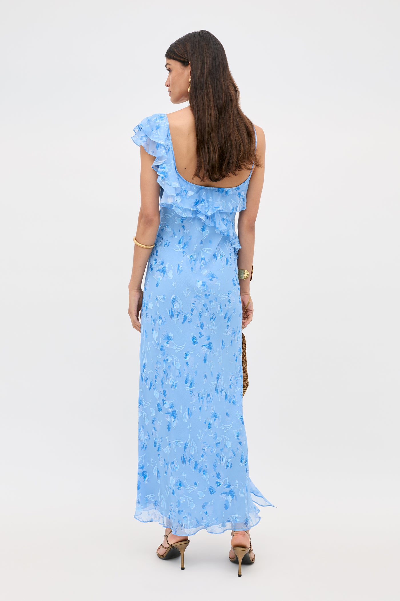 A woman with long brown hair faces away, wearing the Ester - Ava Devore Blue: a light blue, one-shoulder dress with ruffles and a floral pattern. She pairs it with heeled sandals, gold jewelry, and holds a small brown handbag.