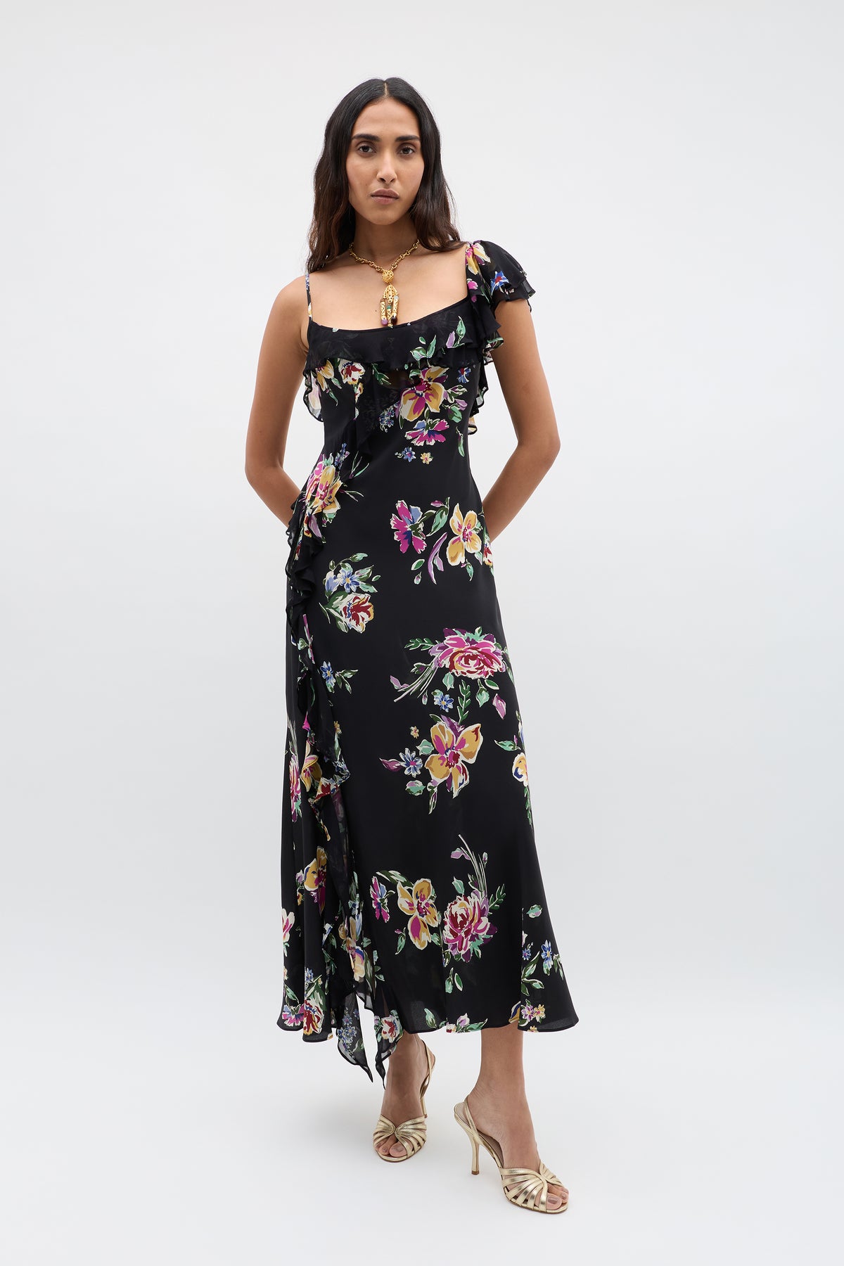 A woman stands against a plain background wearing the Ester - Vintage Bloom Black dress with jacquard florals, ruffled accents, and thin straps, styled with gold heeled sandals and a statement gold necklace.