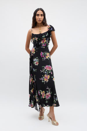 A woman stands against a plain background wearing the Ester - Vintage Bloom Black dress with jacquard florals, ruffled accents, and thin straps, styled with gold heeled sandals and a statement gold necklace.