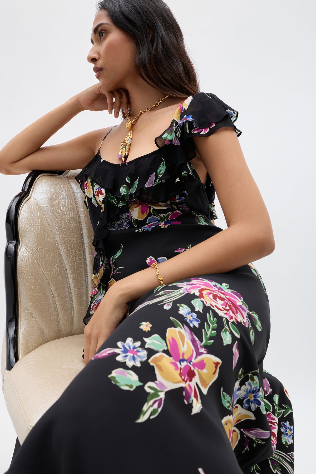 A woman wearing the Ester - Vintage Bloom Black dress with jacquard florals sits sideways on a cream chair. With long dark hair and gold jewelry, she gazes thoughtfully to the side against a plain background.