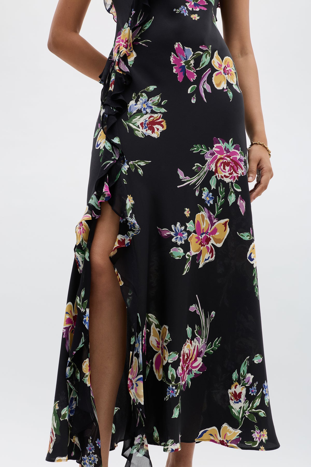 A woman wears the Ester - Vintage Bloom Black dress, featuring black jacquard fabric with pink, yellow, and green floral patterns. A ruffled slit reveals part of her left leg, adding vintage charm against a light background.