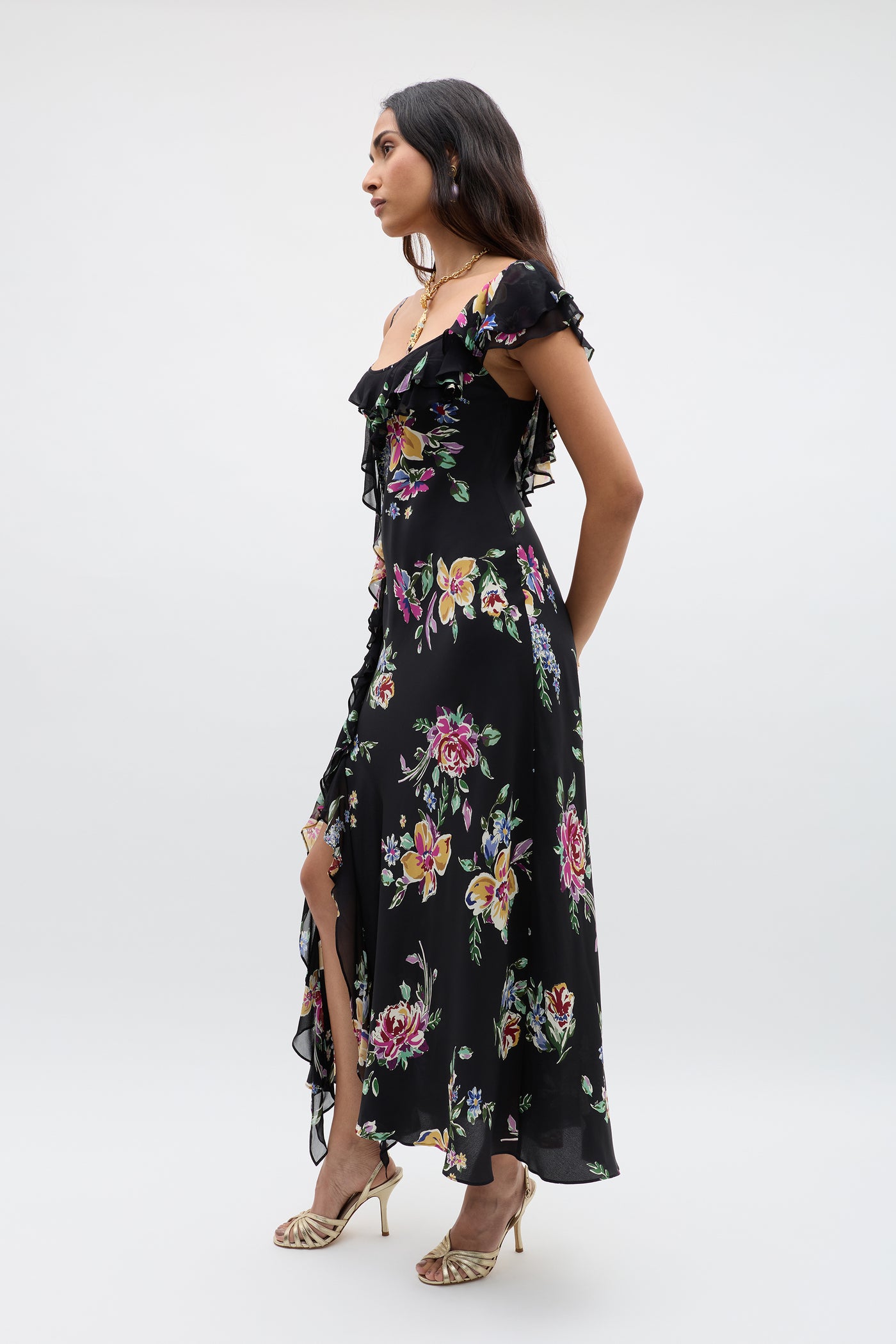 A woman stands sideways in the Ester - Vintage Bloom Black dress, featuring off-shoulder jacquard florals, ruffles, and a thigh-high slit, paired with gold strappy heels against a plain light background.