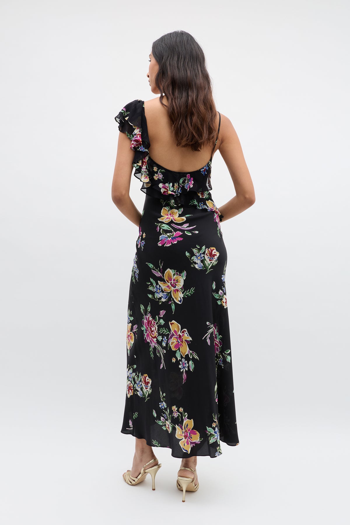 A woman with wavy dark hair faces away in the Ester - Vintage Bloom Black one-shoulder dress with ruffle details and beige heeled sandals, set against a plain light background.