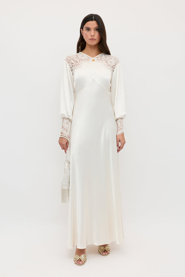 A woman stands facing forward in the Axelle - Pearl, a long white satin dress with lace-detailed shoulders and sleeves. She holds a small white tassel purse, wears light open-toed shoes, and stands against a plain white background.