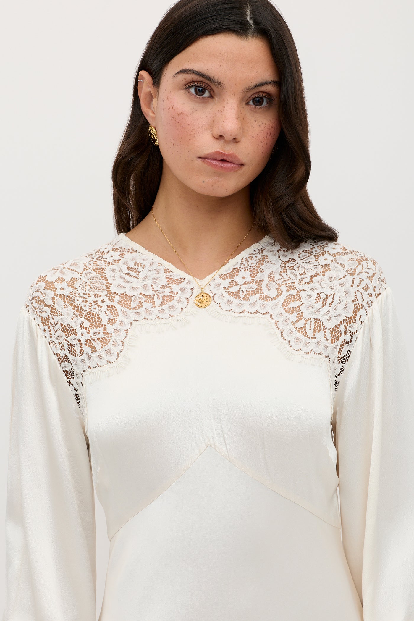 A woman with long dark hair wears the cream Axelle - Pearl dress featuring intricate lace shoulders and a gold necklace. She gazes neutrally at the camera, set against a plain background.