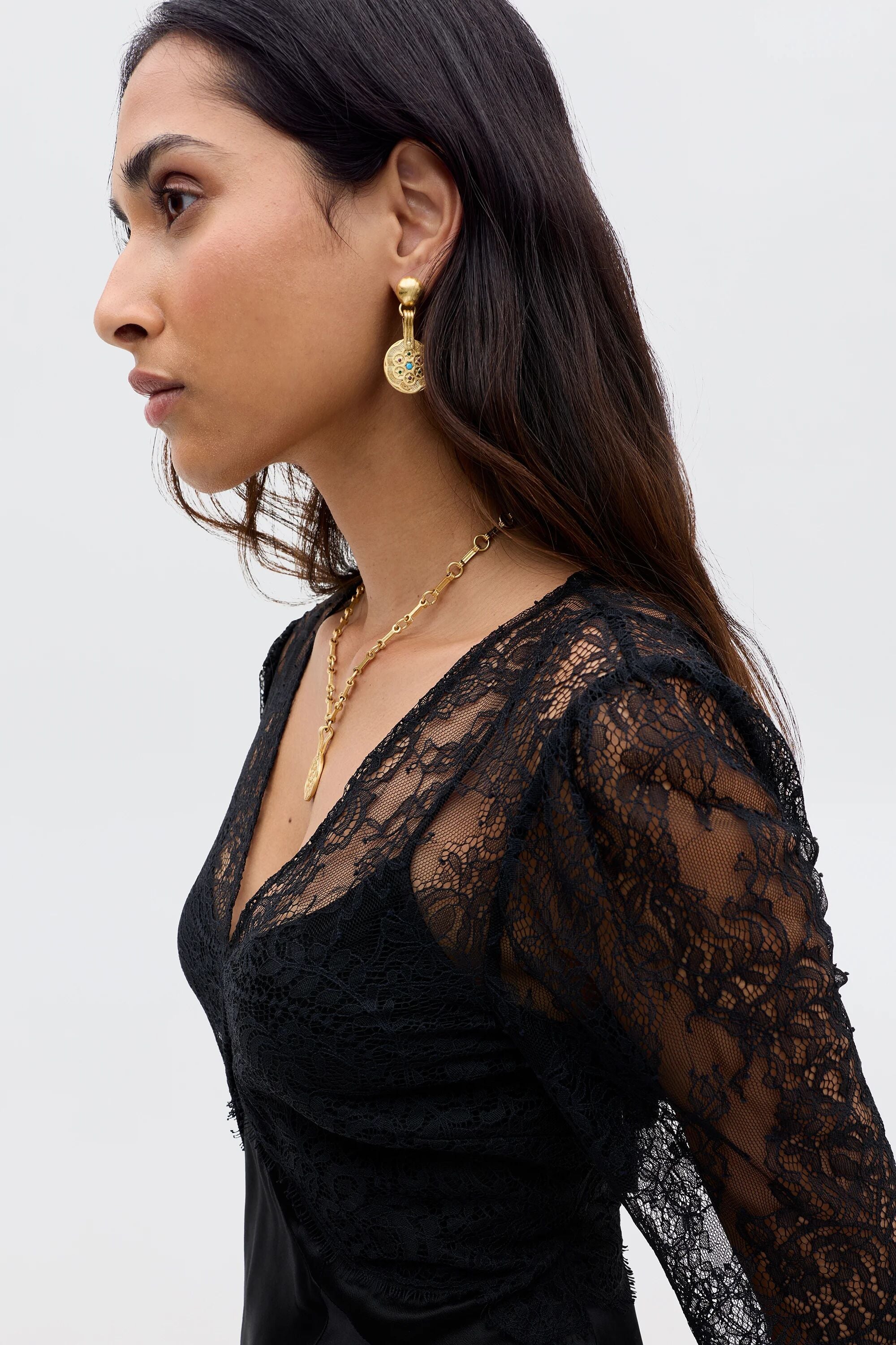 A woman in profile wears the Laoise - Black vintage-inspired dress with black lace, paired with gold drop earrings and a gold pendant necklace. She has long dark hair and a calm expression against a plain light background.