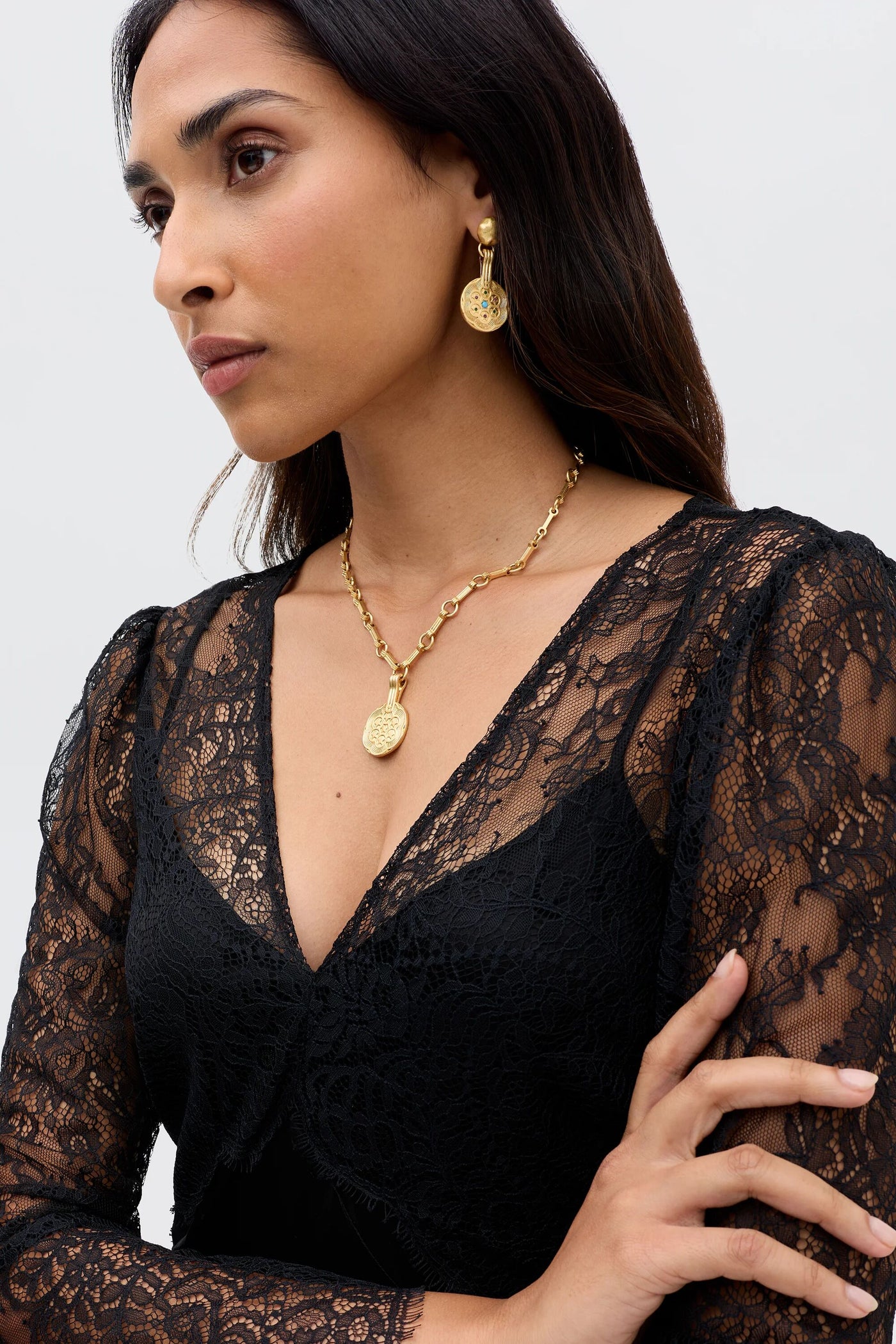 Wearing the Laoise - Black satin and lace dress, a woman poses with arms crossed, gold coin earrings and matching necklace, looking to the side against a plain light background.