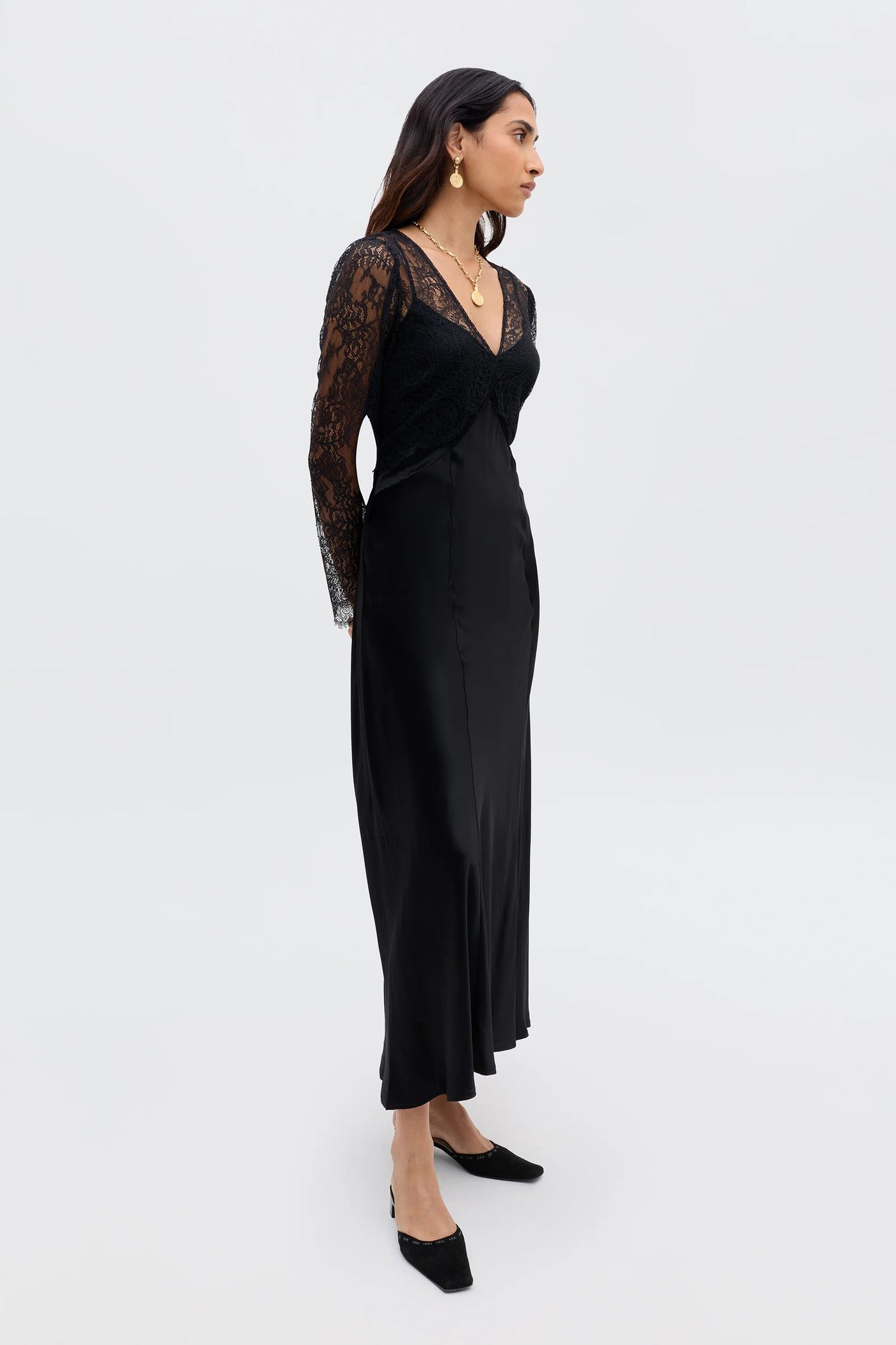 A woman in profile wears the Laoise - Black dress, featuring vintage-inspired satin and lace, long sleeves, a V-neckline, and a silky skirt. She accessorizes with gold earrings, layered necklaces, and black low-heeled shoes against a light backdrop.