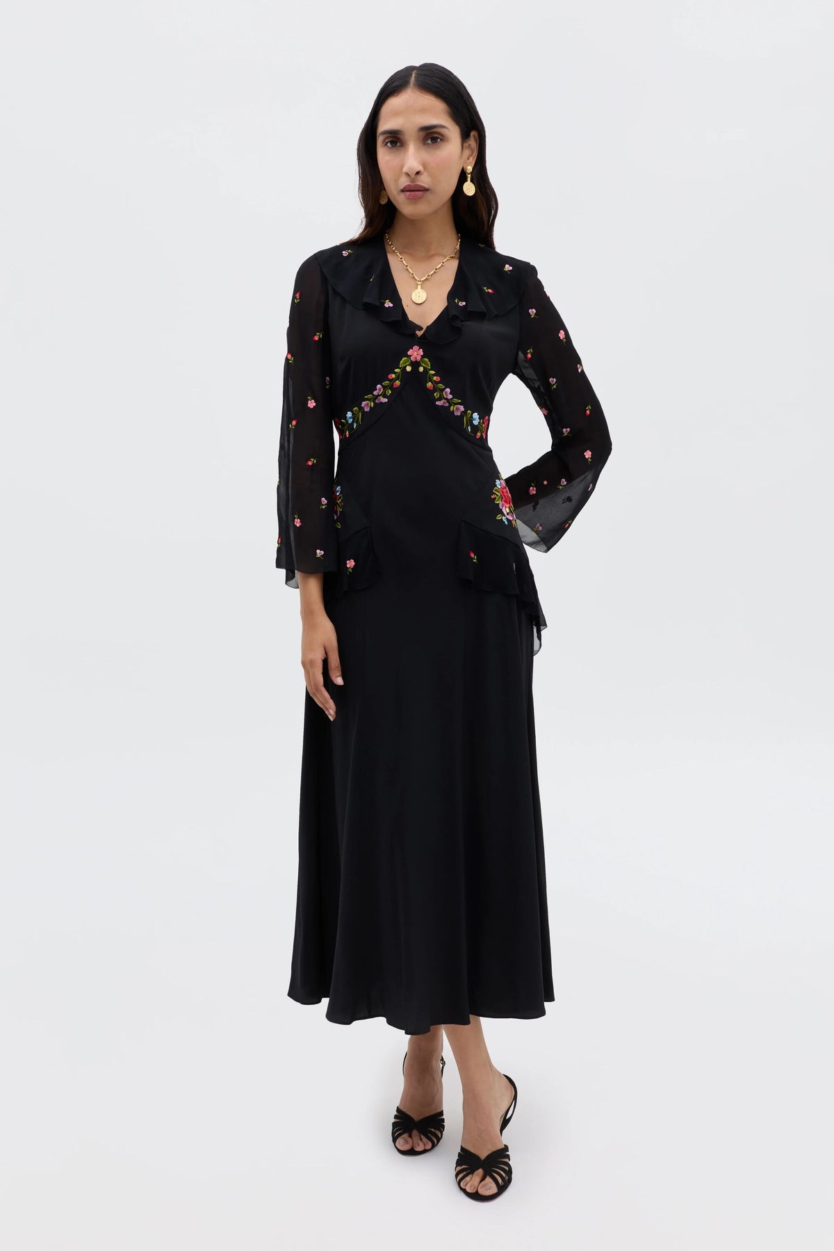 A woman stands against a white background wearing the Cedar - Bouquet Embroidery Black dress, featuring sheer embroidered sleeves with colorful floral details, paired with black heeled sandals and gold jewelry.