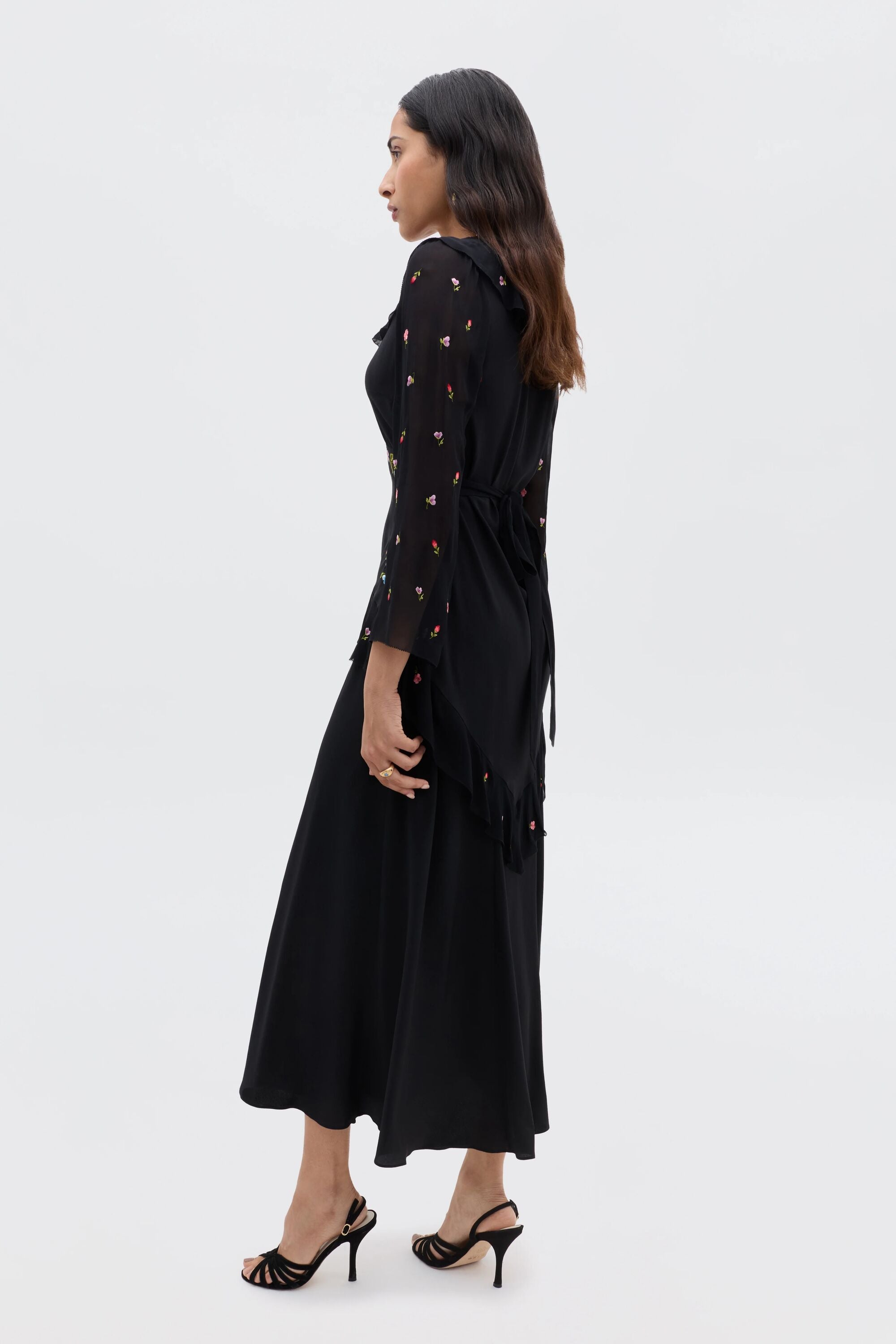 A woman in profile wears the Cedar - Bouquet Embroidery Black dress, featuring subtle bouquet embroidery, a tied waist, and long sleeves. She pairs the long black silk dress with black heeled sandals; her hair is worn loose.