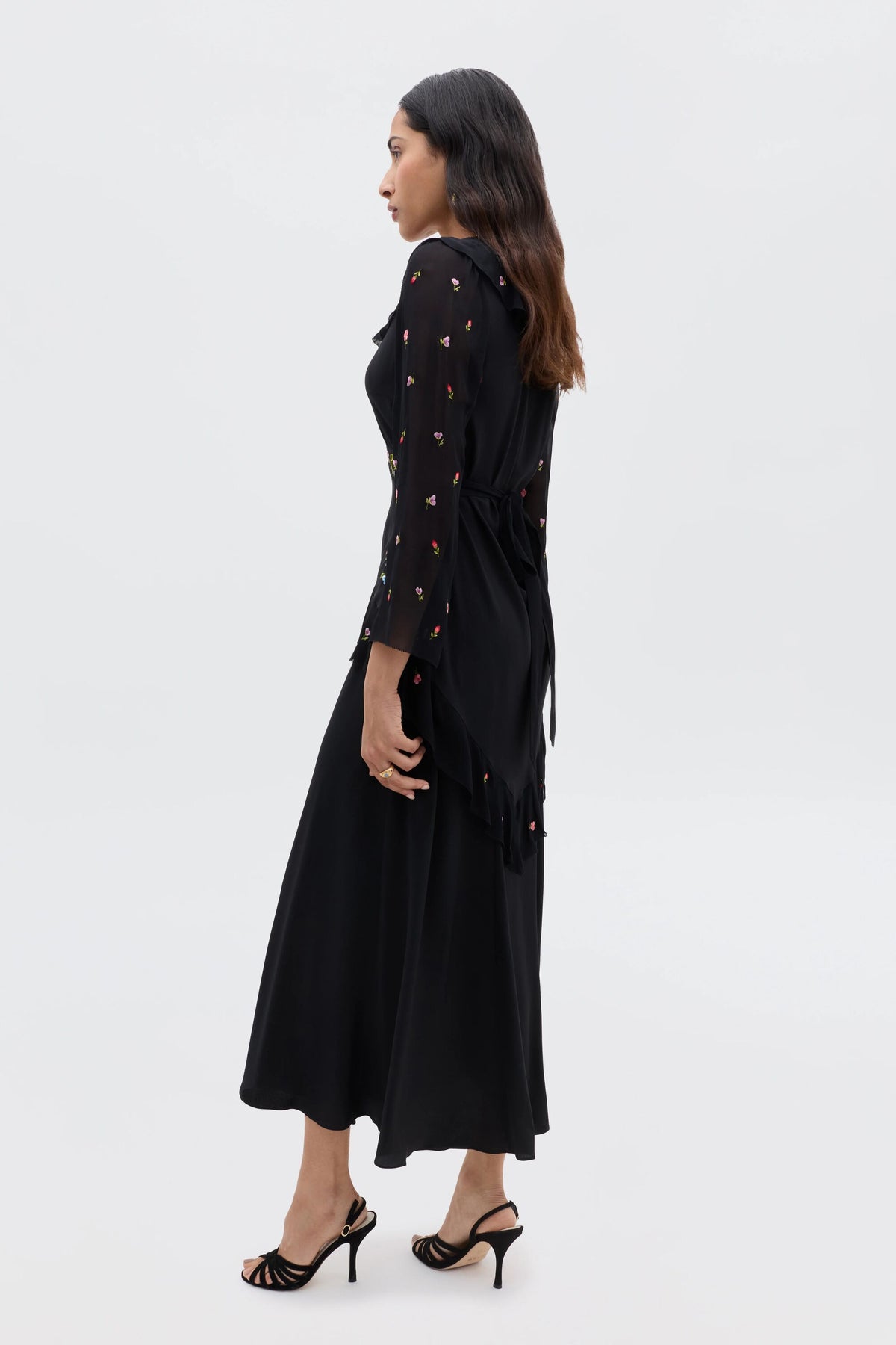 A woman in profile wears the Cedar - Bouquet Embroidery Black dress, featuring subtle bouquet embroidery, a tied waist, and long sleeves. She pairs the long black silk dress with black heeled sandals; her hair is worn loose.