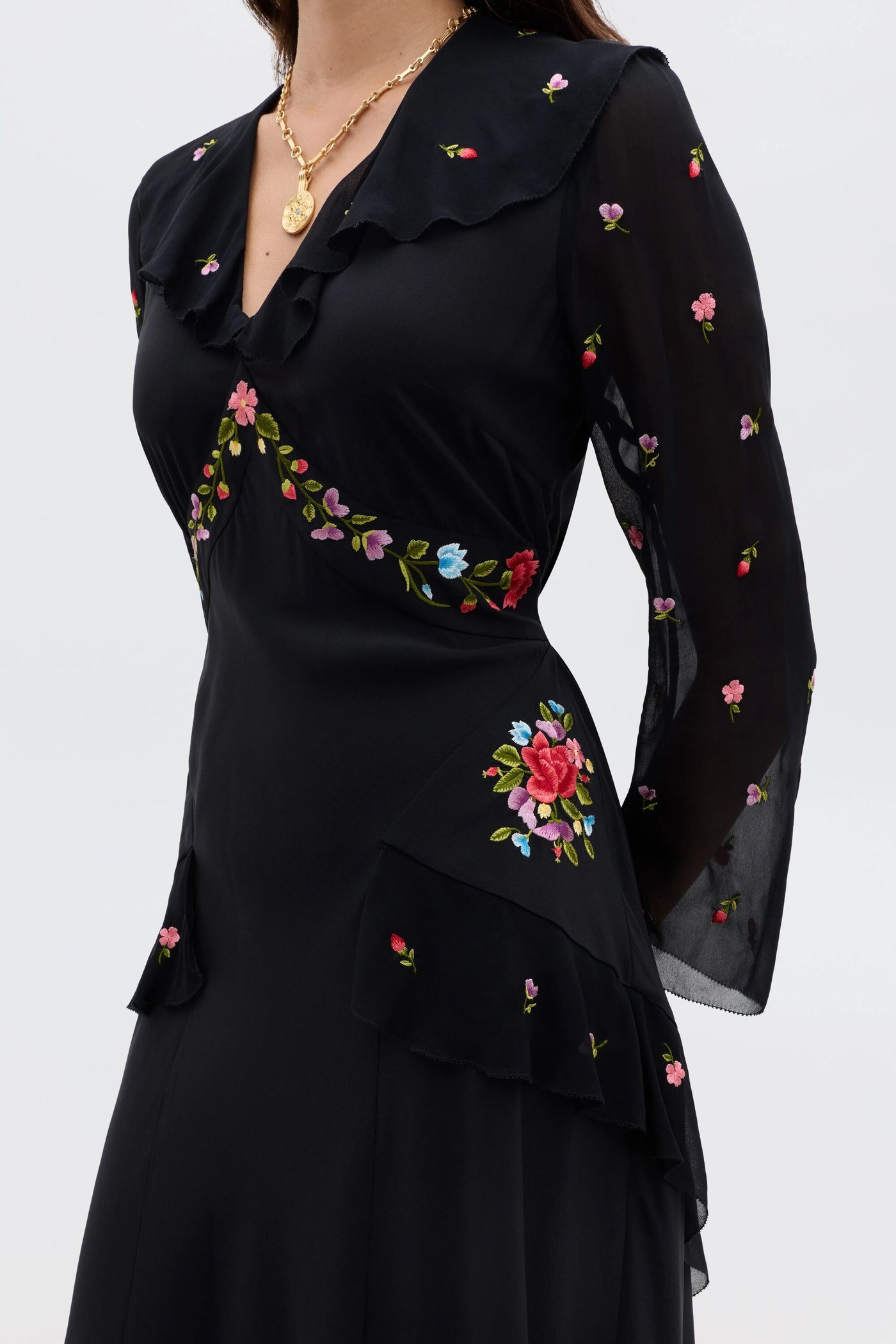 A woman wears the Cedar - Bouquet Embroidery Black dress featuring sheer sleeves, ruffle details, and gold layered necklaces. The image is cropped above her mouth and mid-thigh.