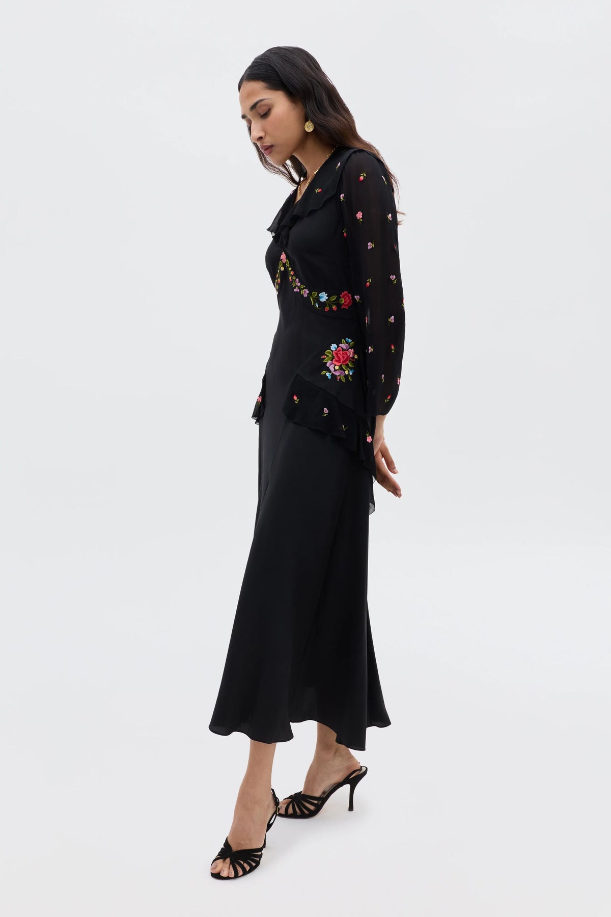 A woman stands in profile wearing the Cedar - Bouquet Embroidery Black dress, featuring intricate floral embroidery on the sleeves, waist, and chest. She has long hair, wears heeled sandals, and gazes downward thoughtfully.