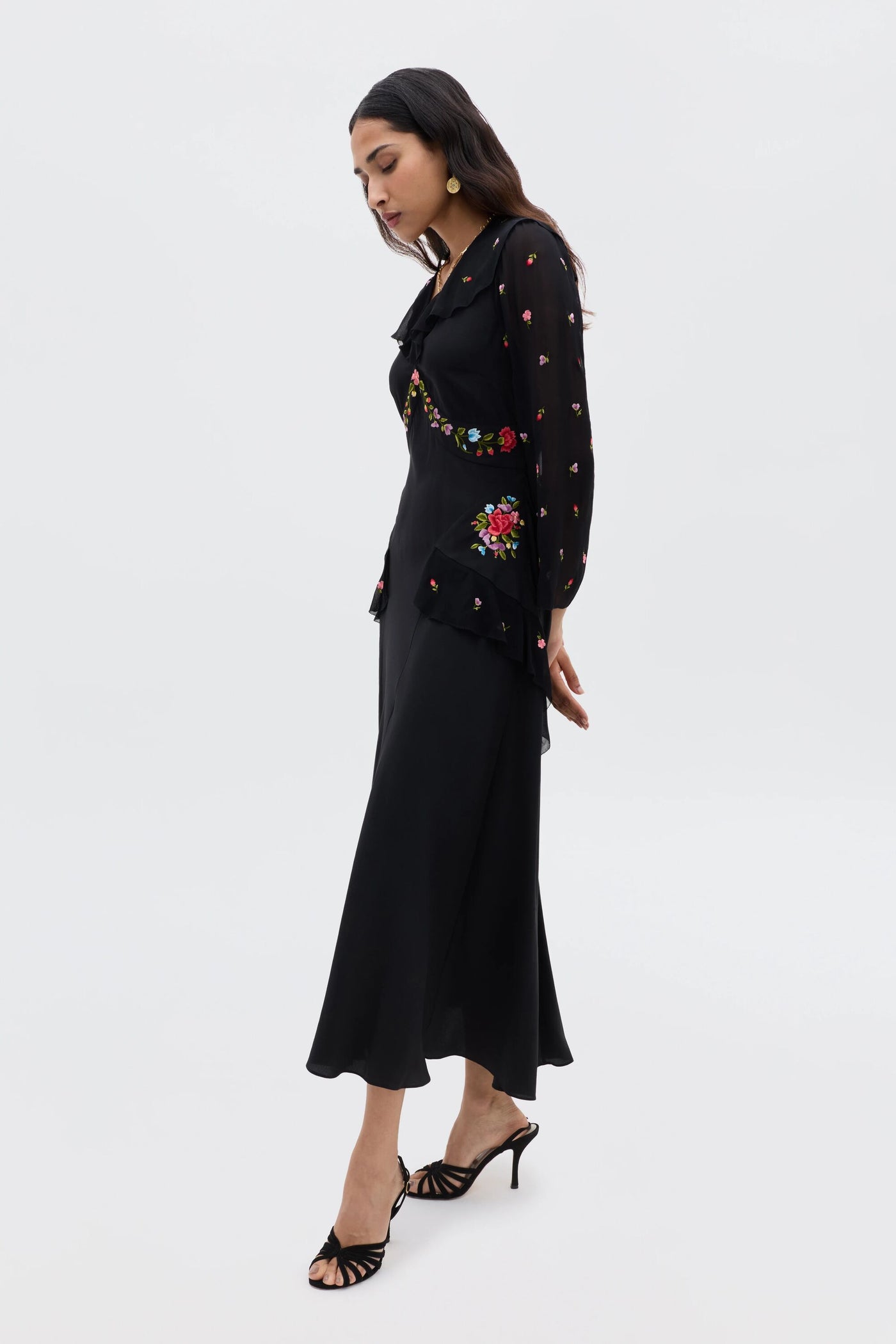 A woman stands in profile wearing the Cedar - Bouquet Embroidery Black dress, featuring intricate floral embroidery on the sleeves, waist, and chest. She has long hair, wears heeled sandals, and gazes downward thoughtfully.