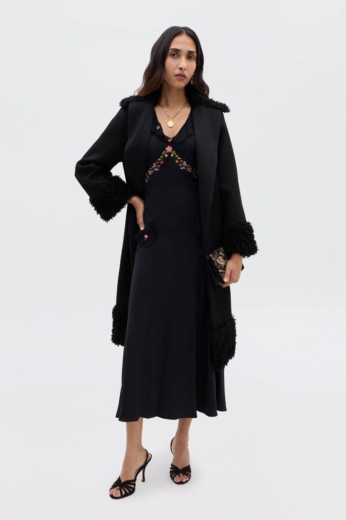 A woman with long dark hair stands against a white background wearing the Cedar - Bouquet Embroidery Black midi silk dress and a long black coat with textured cuffs and collar. She pairs it with black heels and holds a small clutch.