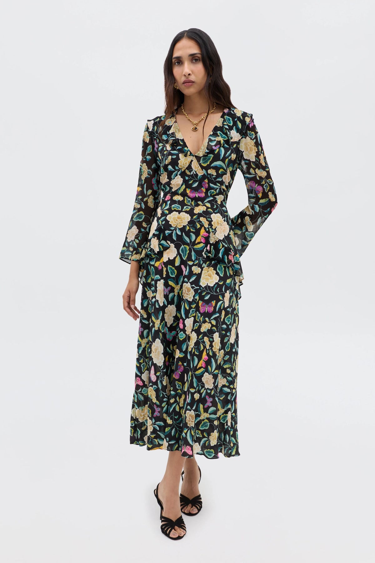 A woman with long, wavy dark hair wears the Cedar - Butterfly Floral Charcoal dress—a long-sleeve, V-neck style with an empire waist and colorful butterfly floral print—paired with black strappy heels and layered gold necklaces.