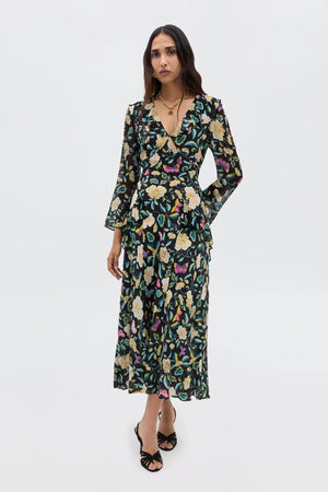 A woman with long, wavy dark hair wears the Cedar - Butterfly Floral Charcoal dress—a long-sleeve, V-neck style with an empire waist and colorful butterfly floral print—paired with black strappy heels and layered gold necklaces.
