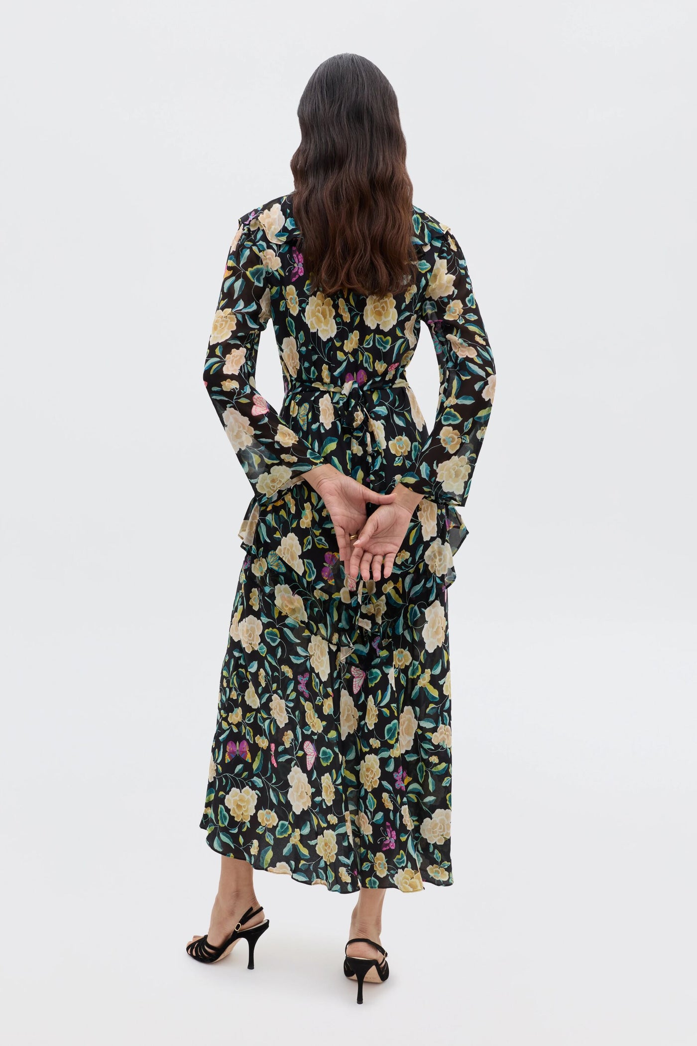 A woman, seen from behind with hands clasped, wears the Cedar - Butterfly Floral Charcoal dress featuring an empire waist and yellow-green florals, paired with black high-heeled sandals against a plain white background.