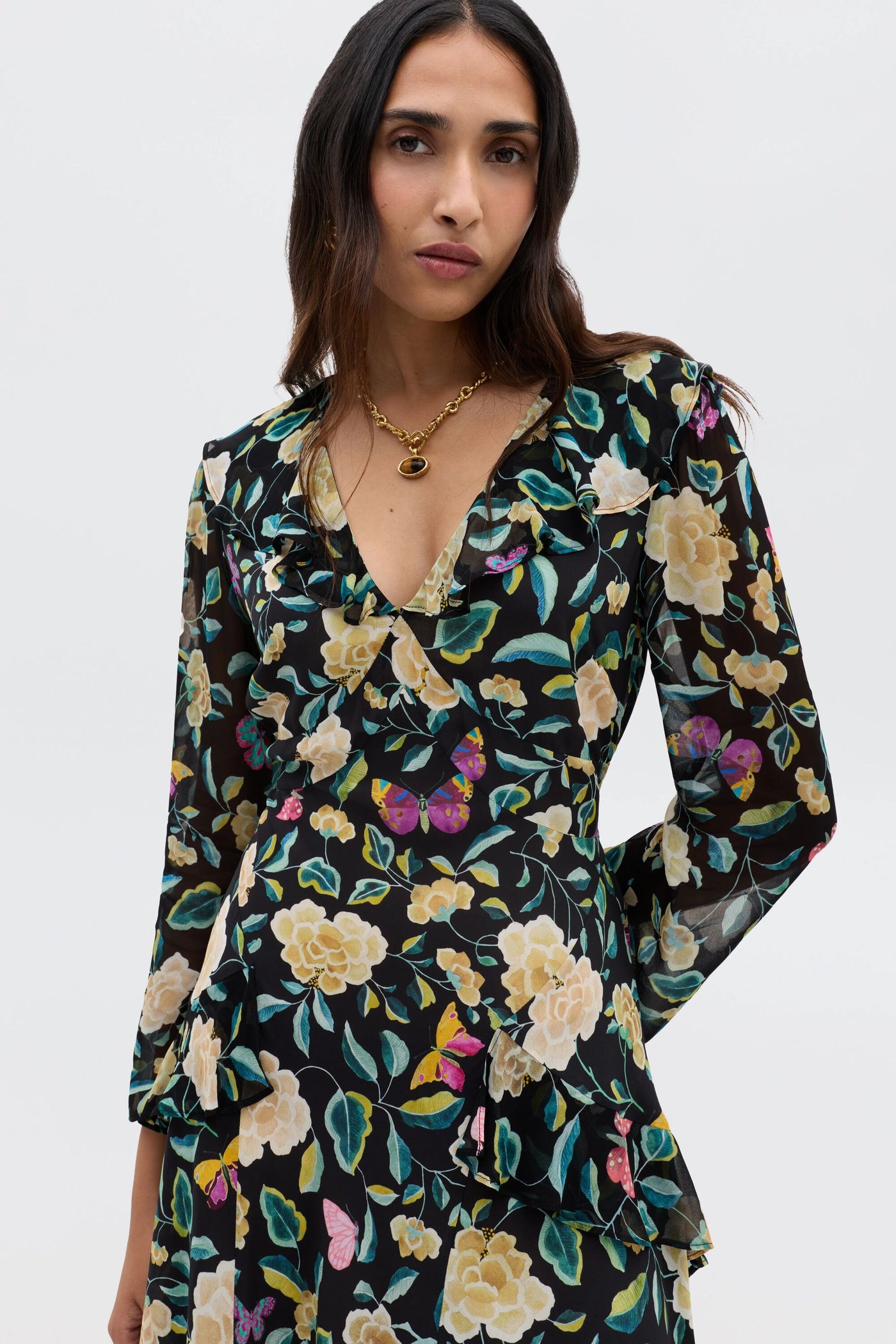 A woman with long dark hair models the Cedar - Butterfly Floral Charcoal dress with an empire waist and a gold necklace, standing against a plain light background and looking confidently at the camera.