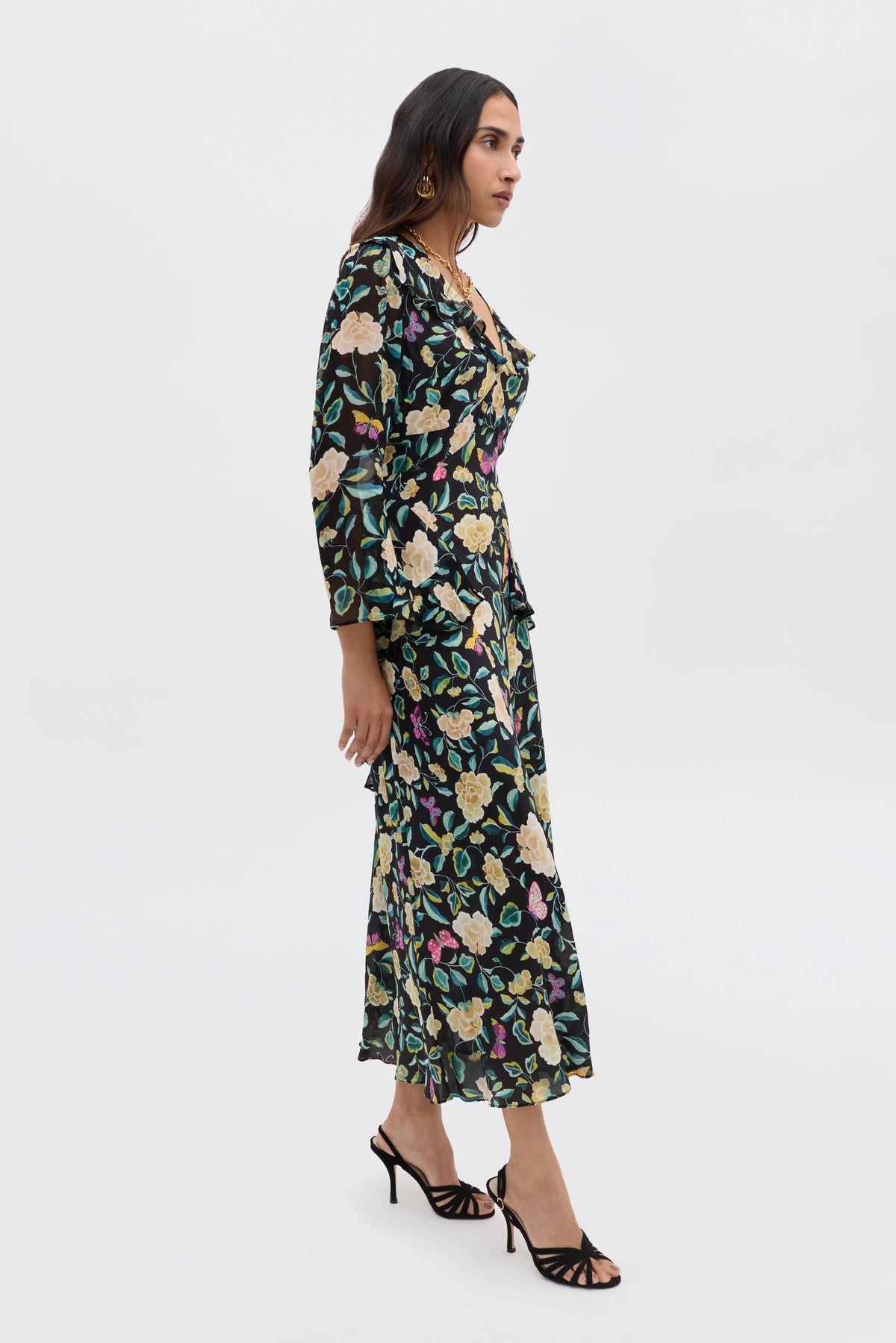 Woman standing sideways wearing the Cedar - Butterfly Floral Charcoal dress, featuring an empire waist and long sleeves, paired with black strappy high-heeled sandals, against a plain white background.