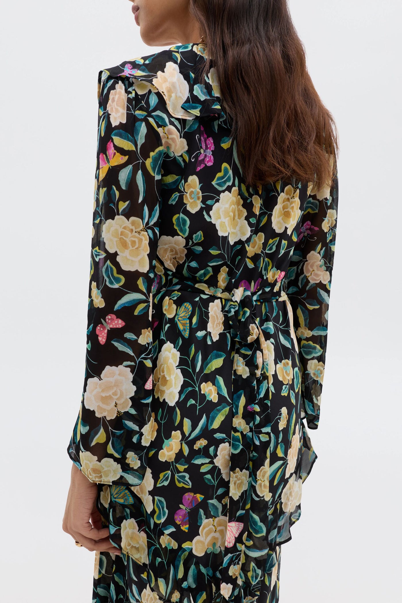 A woman is seen from the back wearing the Cedar - Butterfly Floral Charcoal dress, a black silk piece with a colorful print, featuring an empire waist, waist tie, and elegant long sleeves.