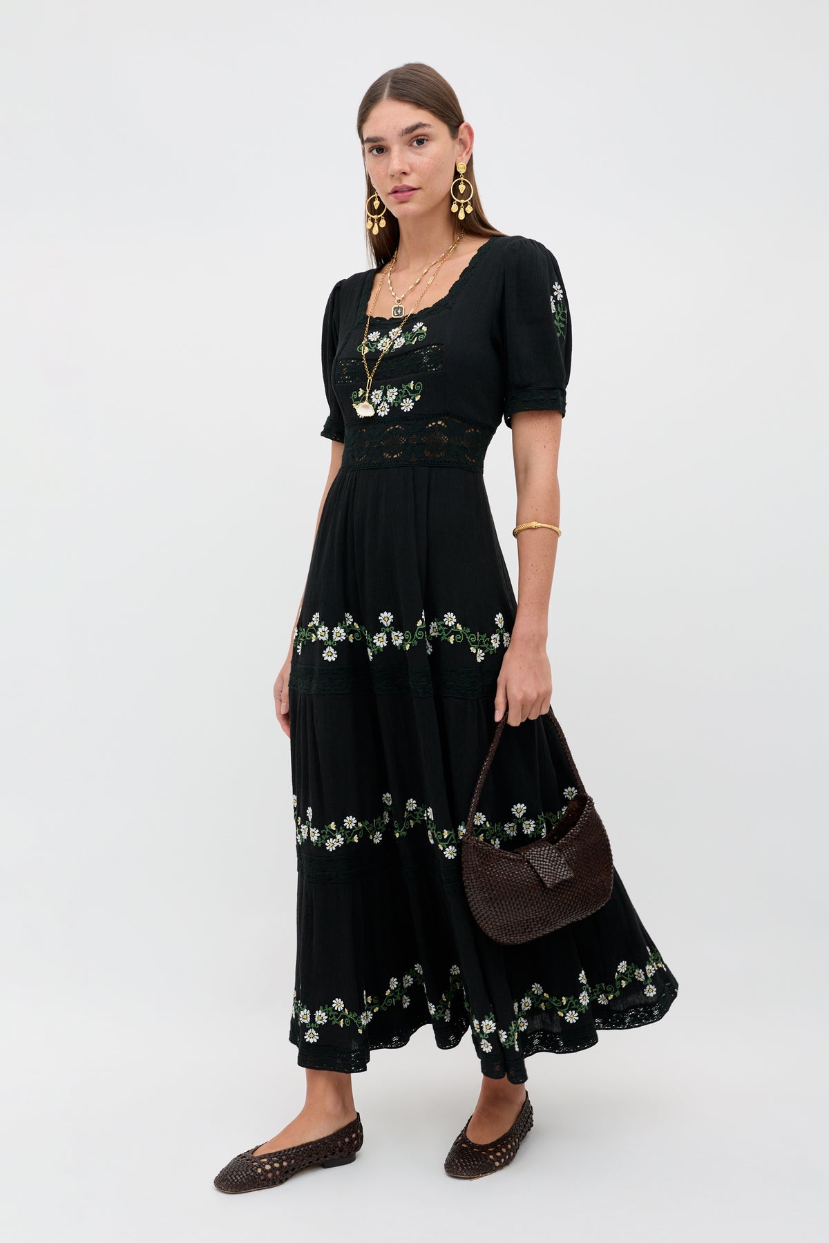 A woman with straight brown hair and a neutral expression stands against a plain background wearing the Philippa - Lilia Embroidery Black ankle-length dress, gold jewelry, brown woven flats, and holding a dark woven handbag.